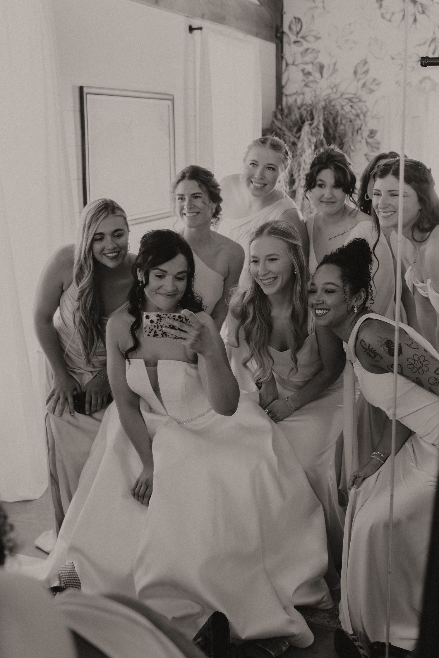 Black and white photo of a smiling bride surrounded by bridesmaids in a bright wedding getting-ready room.