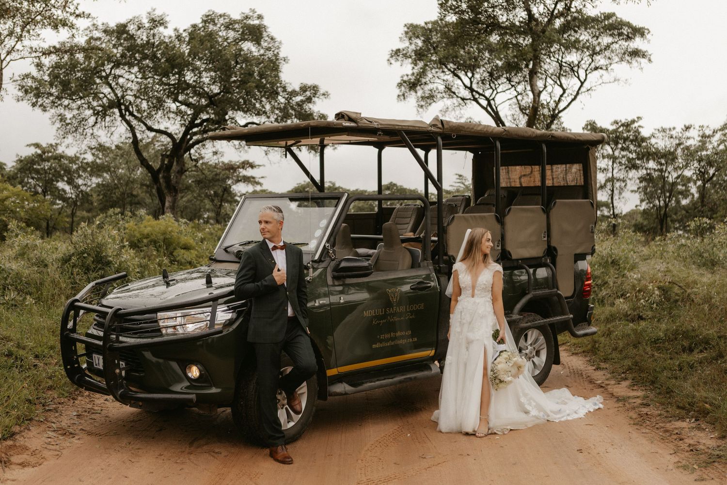 Bride and groom posing beside a dark green safari truck on a dirt road surrounded by African bush trees.
