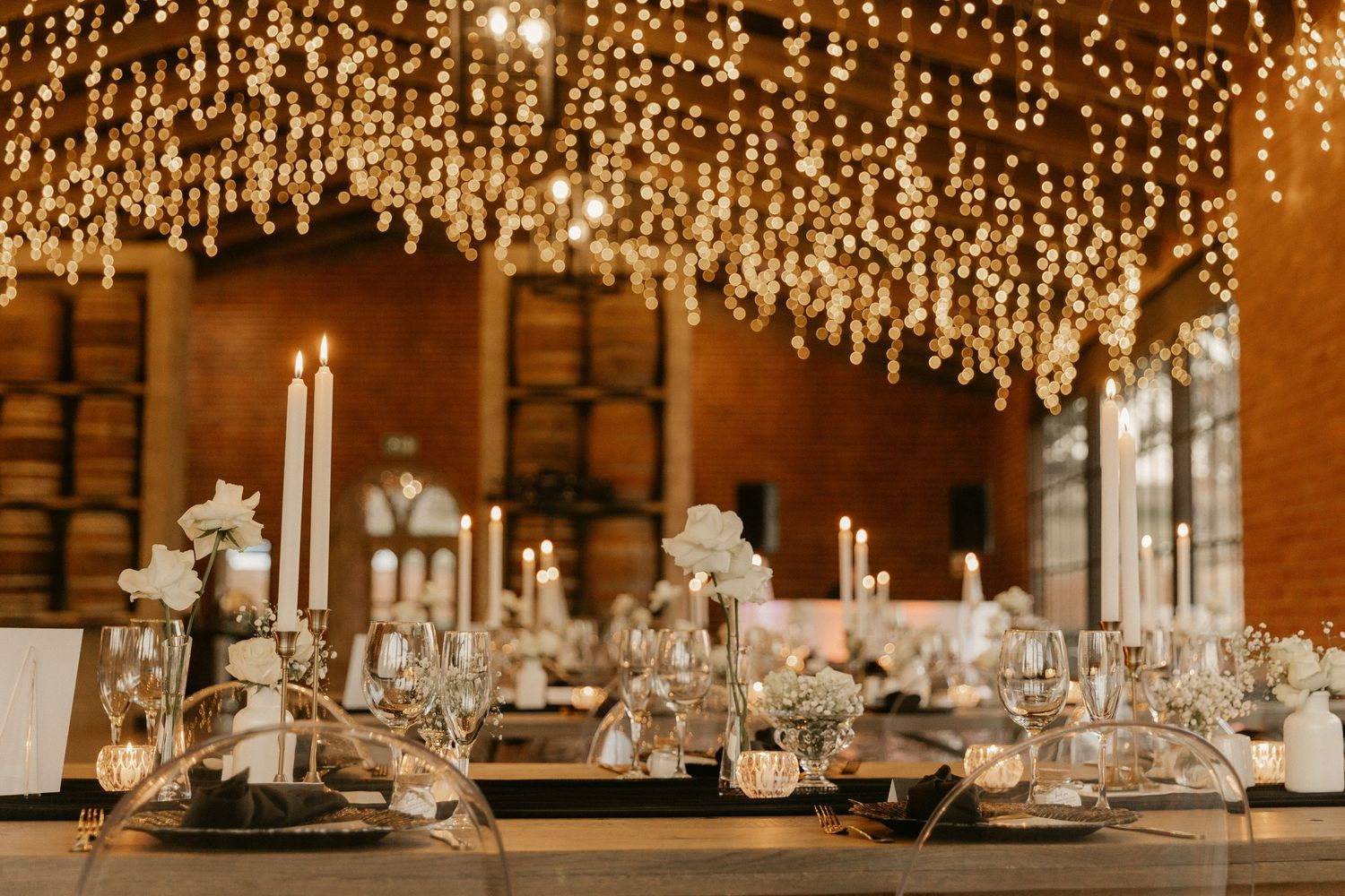 Elegant wedding reception table setting with hanging string lights and white floral centerpieces in an industrial venue.
