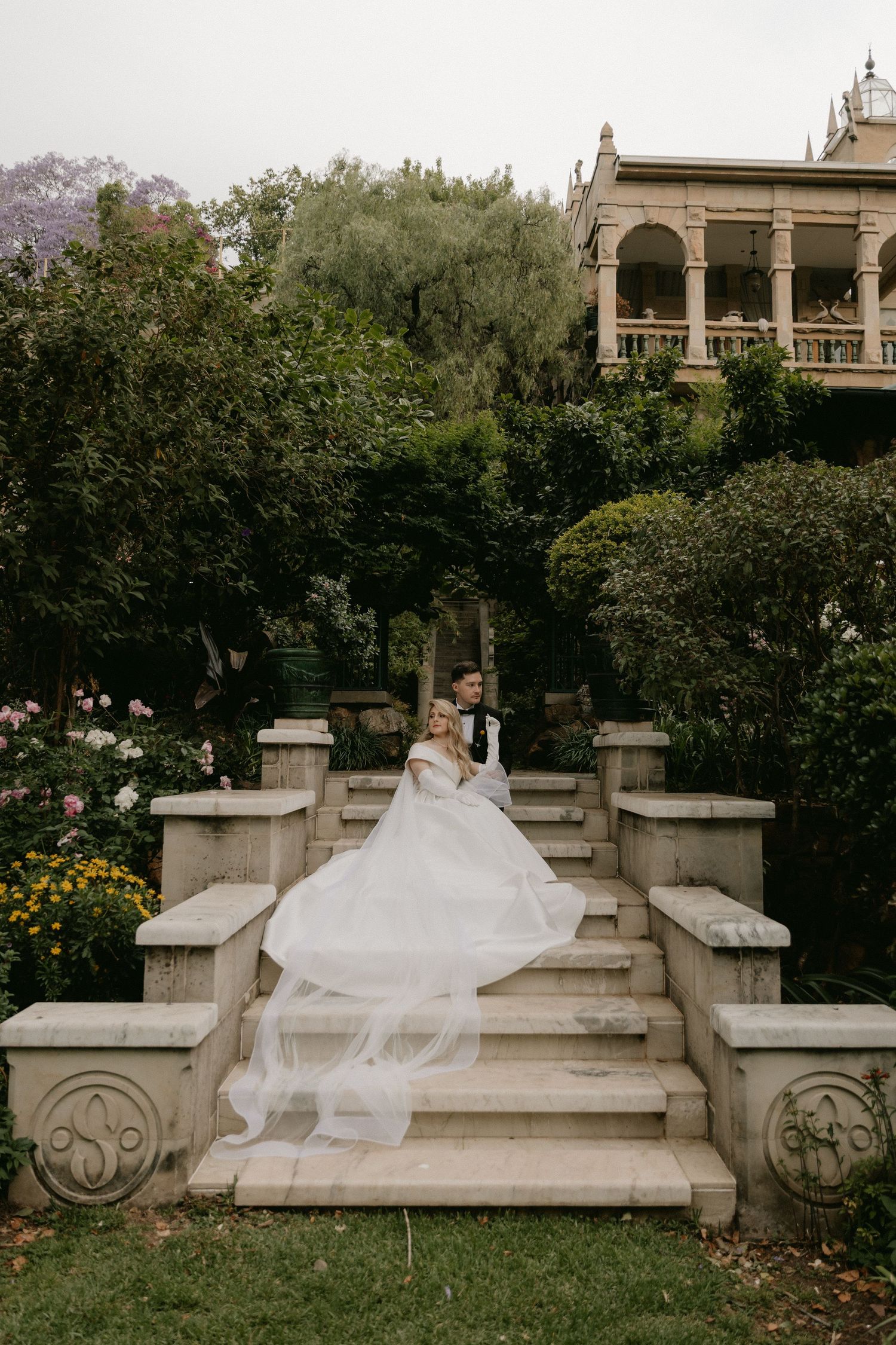 A bride in a flowing white gown poses gracefully on stone garden steps in a romantic outdoor setting.