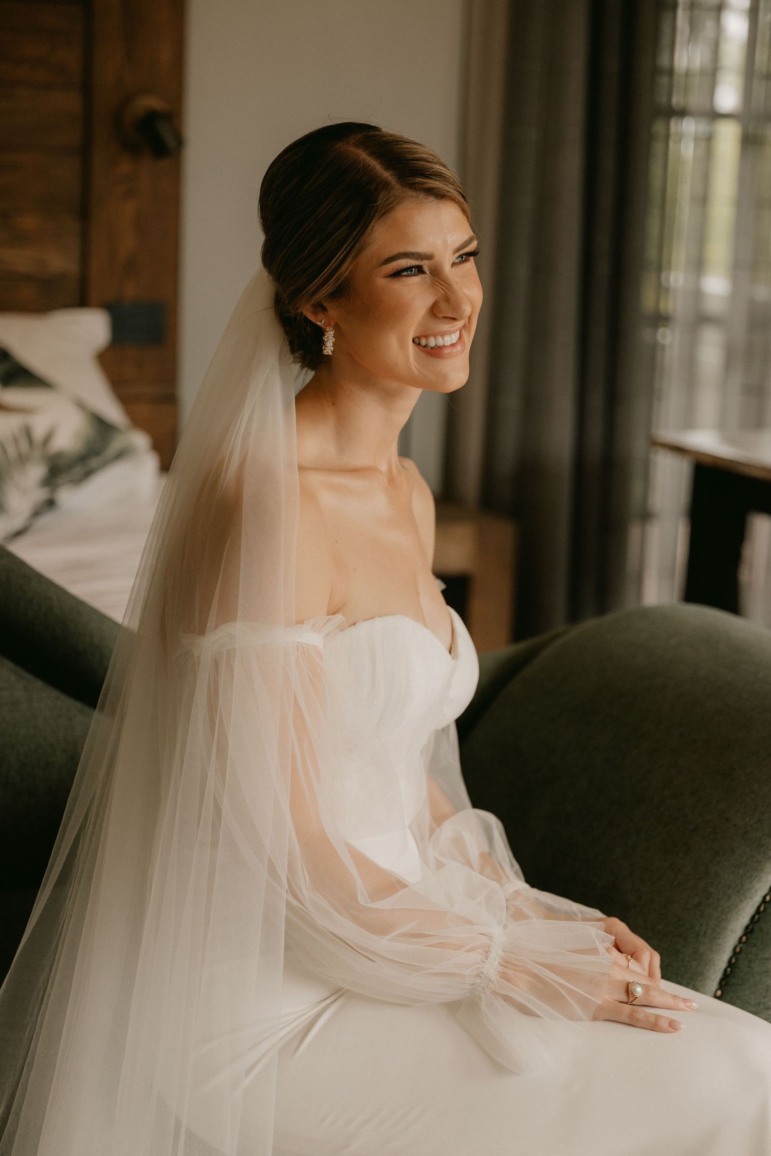 Beautiful bride in white strapless gown with flowing veil smiles joyfully while seated by a window on her wedding day.