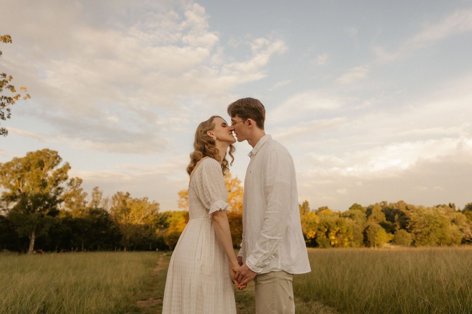 A romantic couple embraces in a golden sunset field with tall grass and warm autumn light glowing around them.