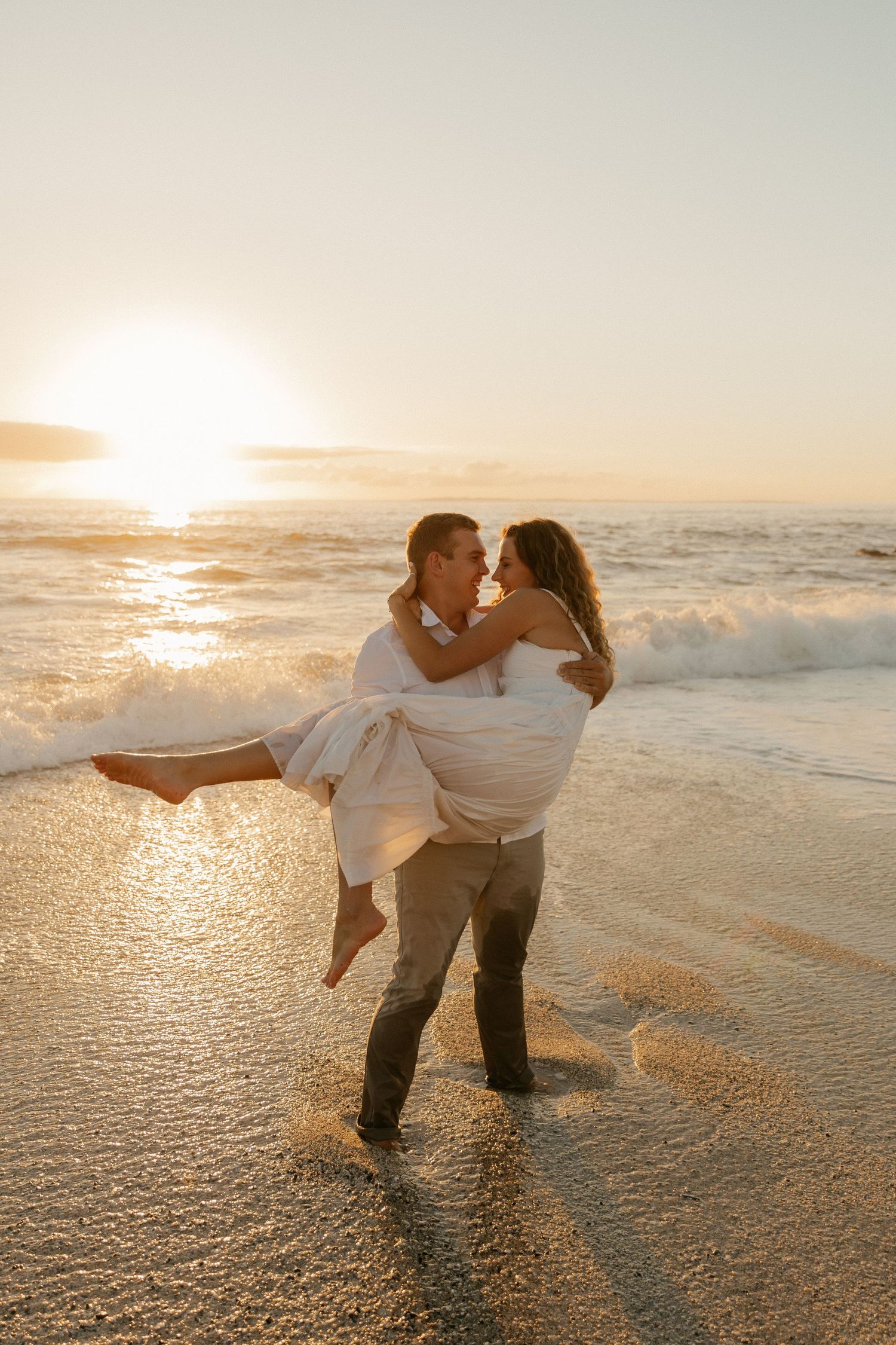 A romantic beach scene with someone lifting their partner in white dress during sunset.