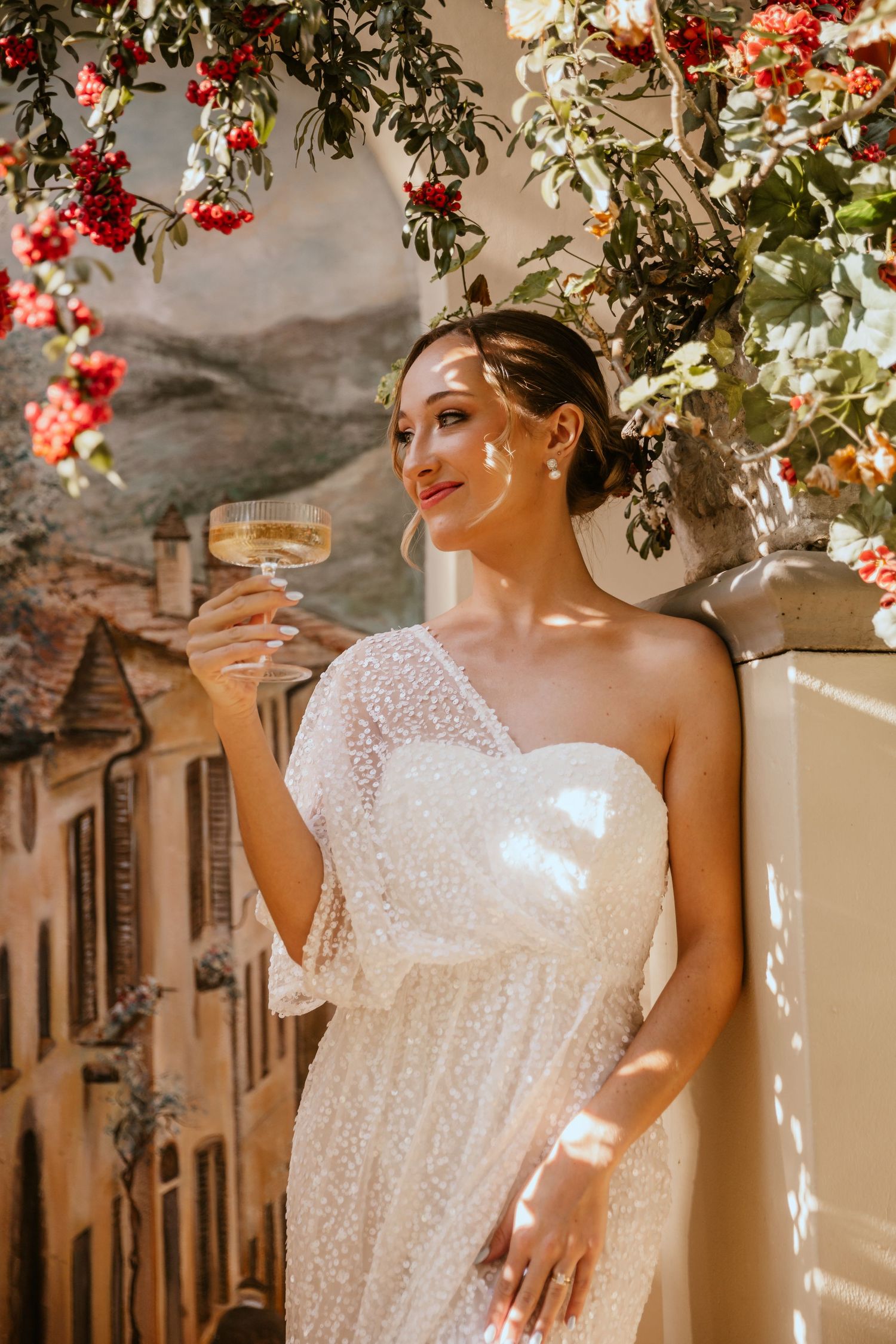 Bride in white dress holding champagne glass under flowering red bougainvillea.