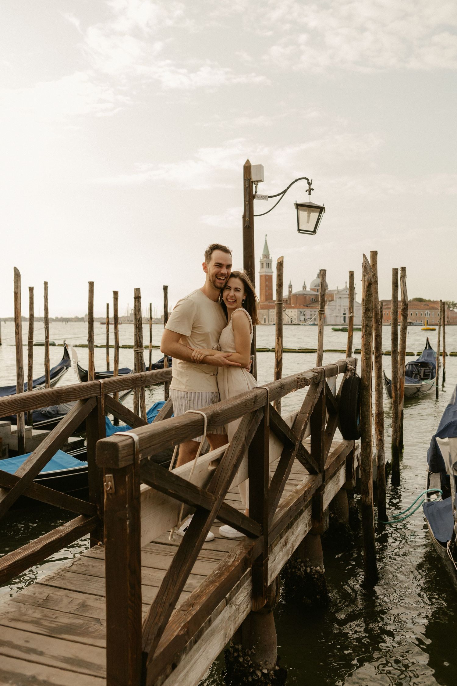 Couple embraces on wooden dock overlooking gondolas and waterfront in Venice, Italy with San Giorgio Maggiore in background.
