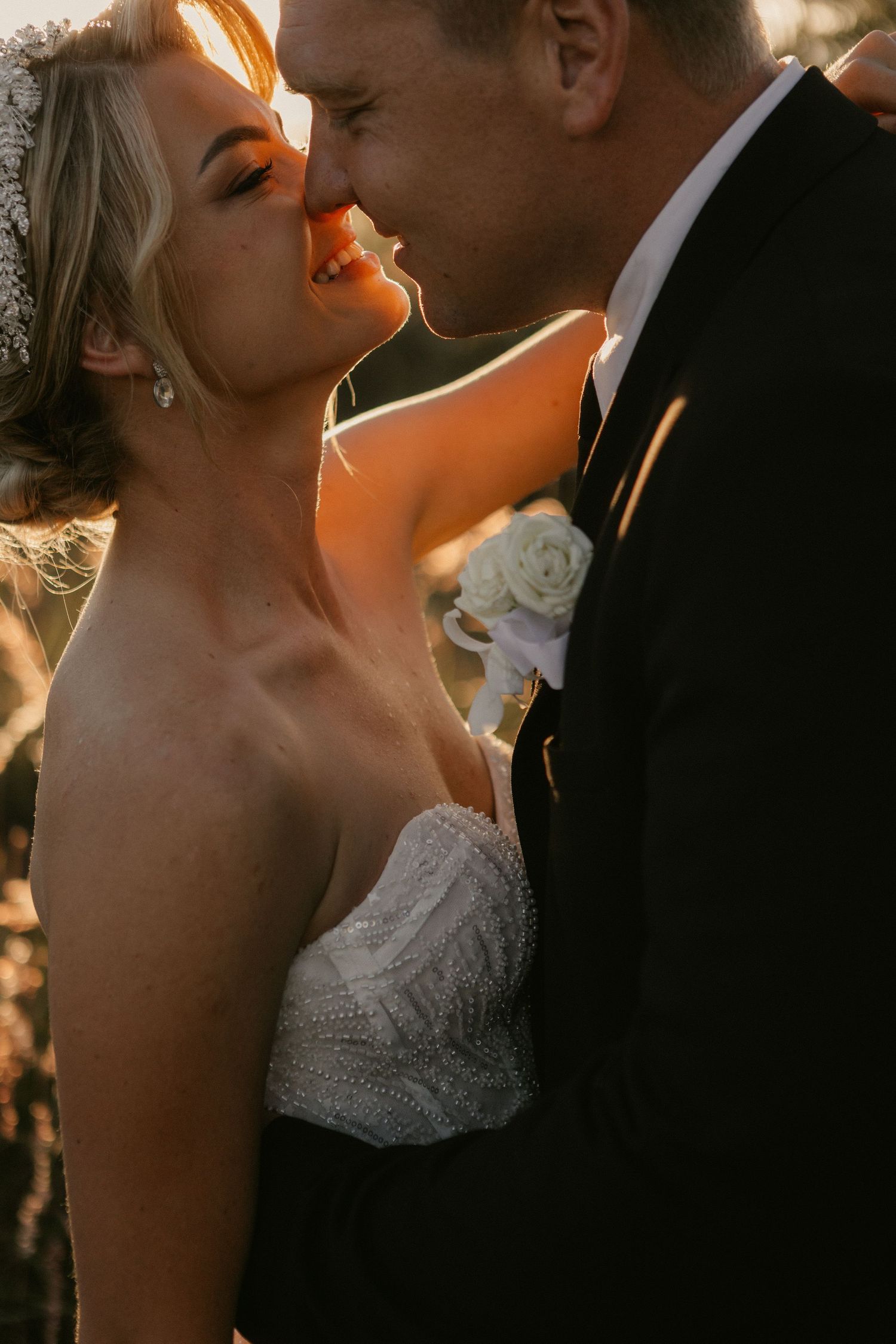 Bride and groom share a romantic golden hour kiss, with bride in strapless lace gown and groom in black suit with white boutonniere.
