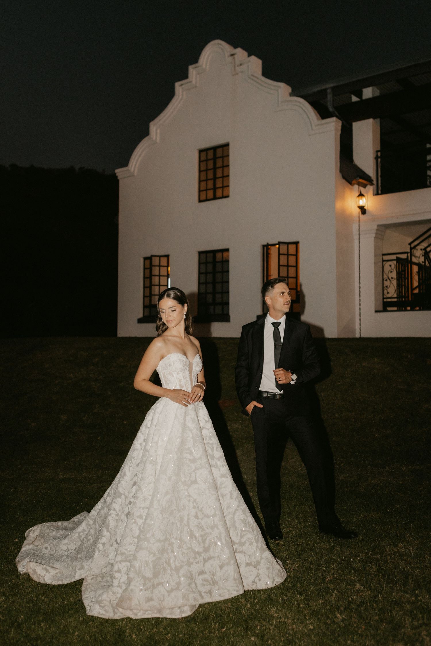 Bride in white ballgown and groom in black tuxedo pose at night outside elegant white Cape Dutch manor.