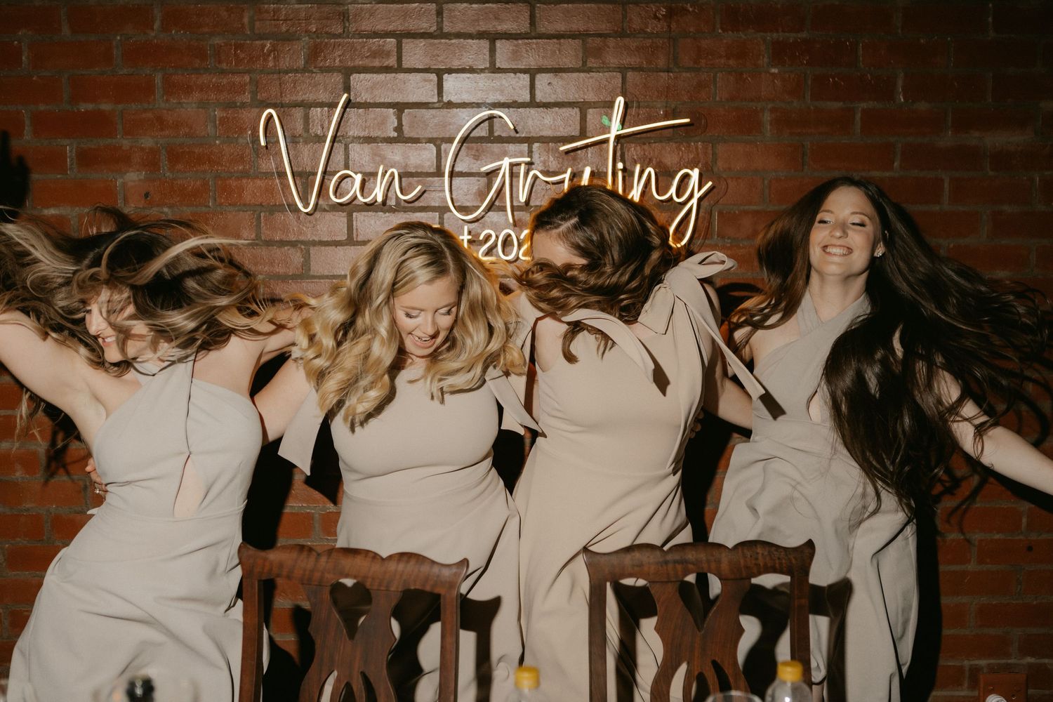 Bridesmaids in champagne dresses dancing under Van Gruting neon sign on brick wall.
