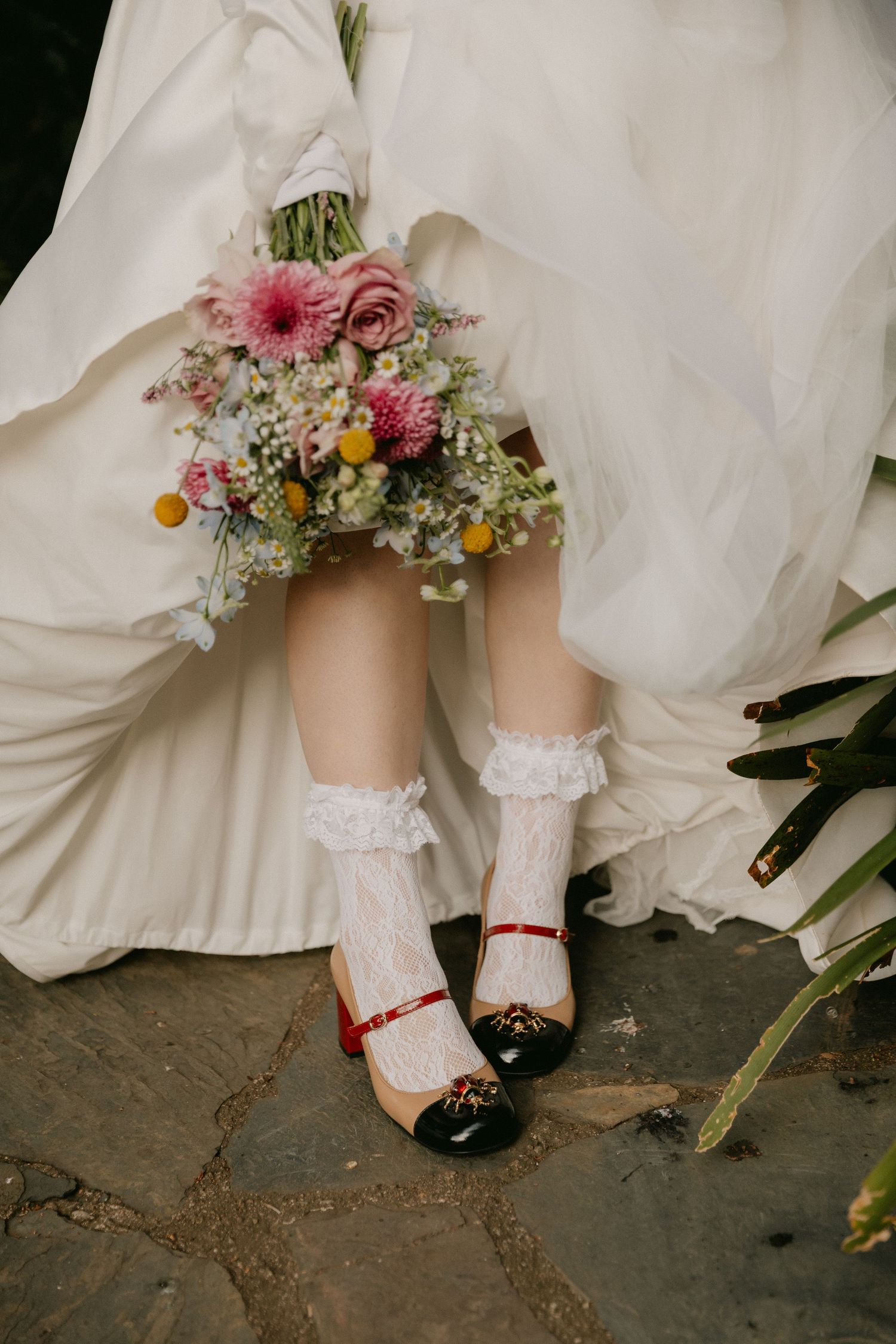 A pair of feet wearing lace socks and black Mary Jane shoes with red straps, surrounded by a white dress and flowers.