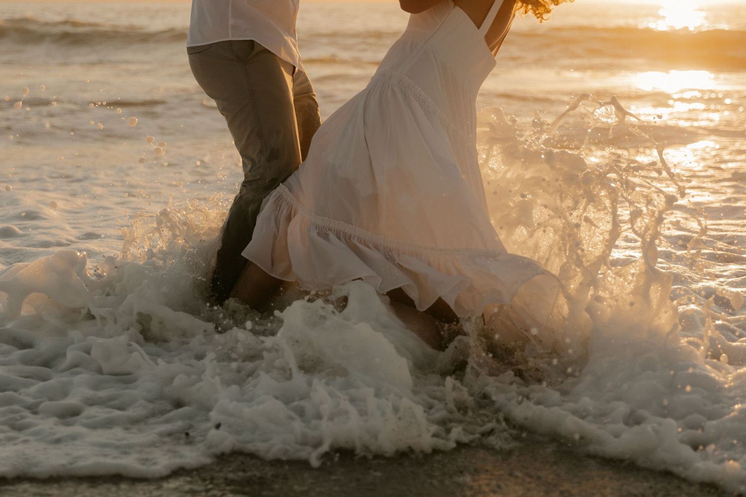 Waves crash around a couple's feet during a romantic beach photoshoot at golden hour.
