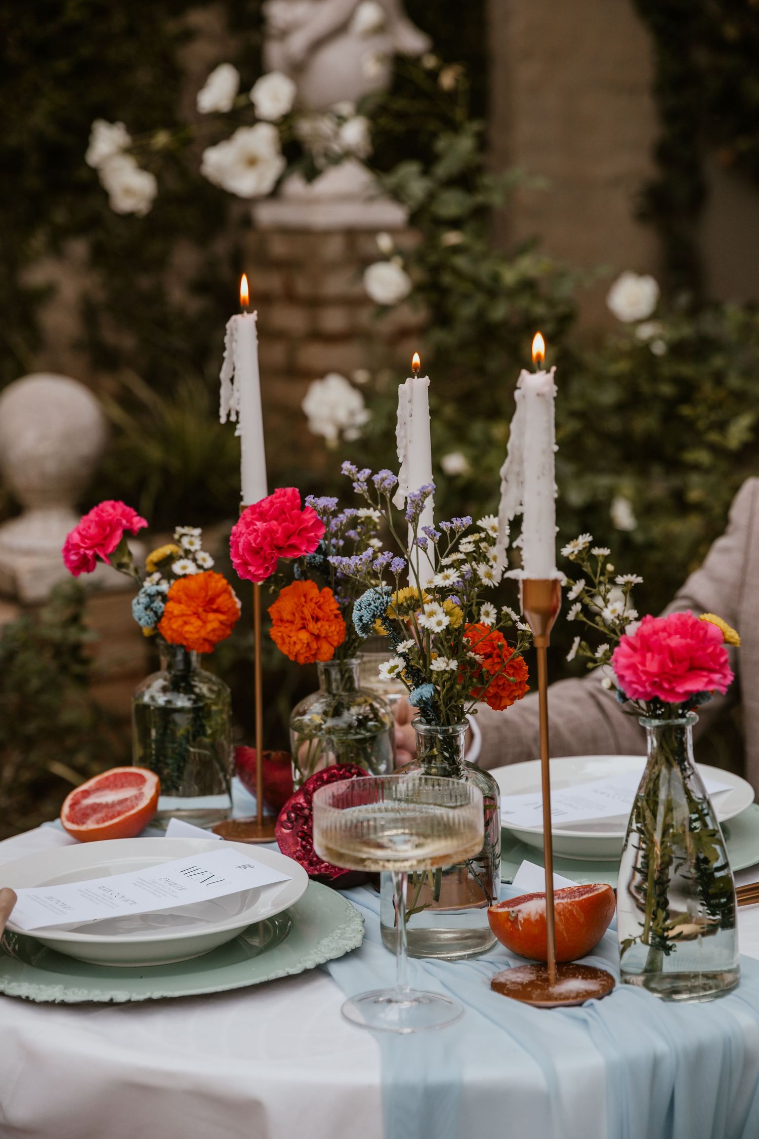 Romantic table setting with white candles, colorful flowers, and grapefruit halves on white tablecloth.