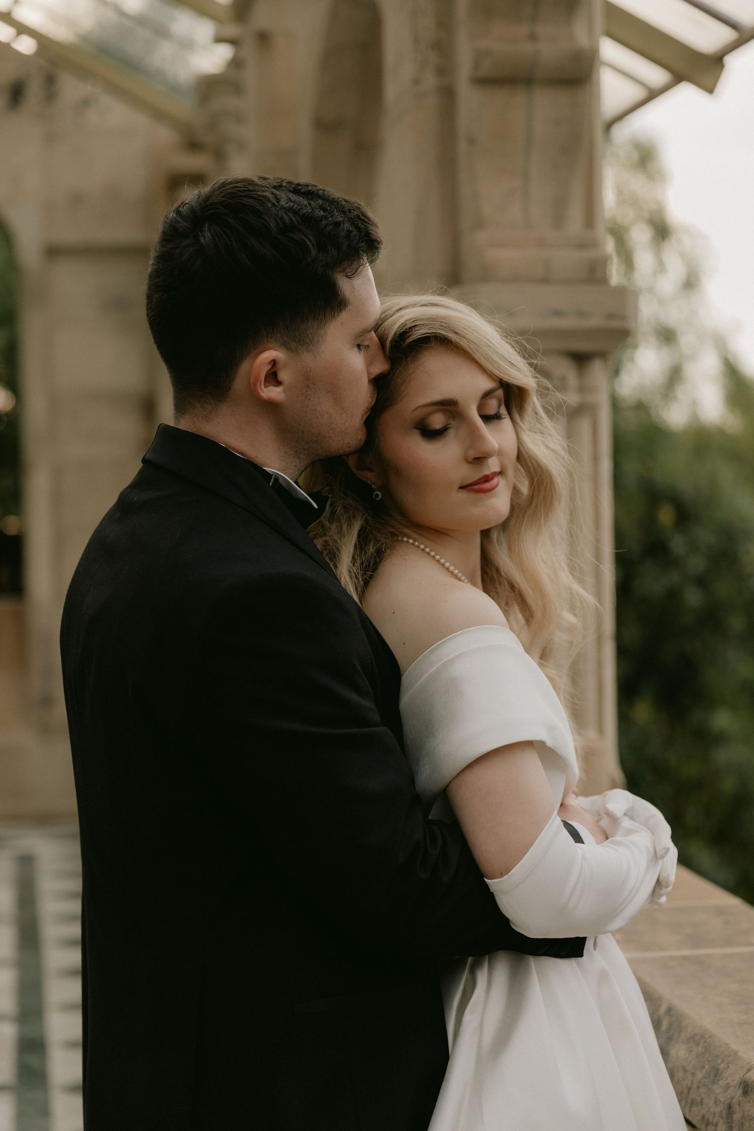 A romantic wedding couple embraces in an elegant stone archway during golden hour.
