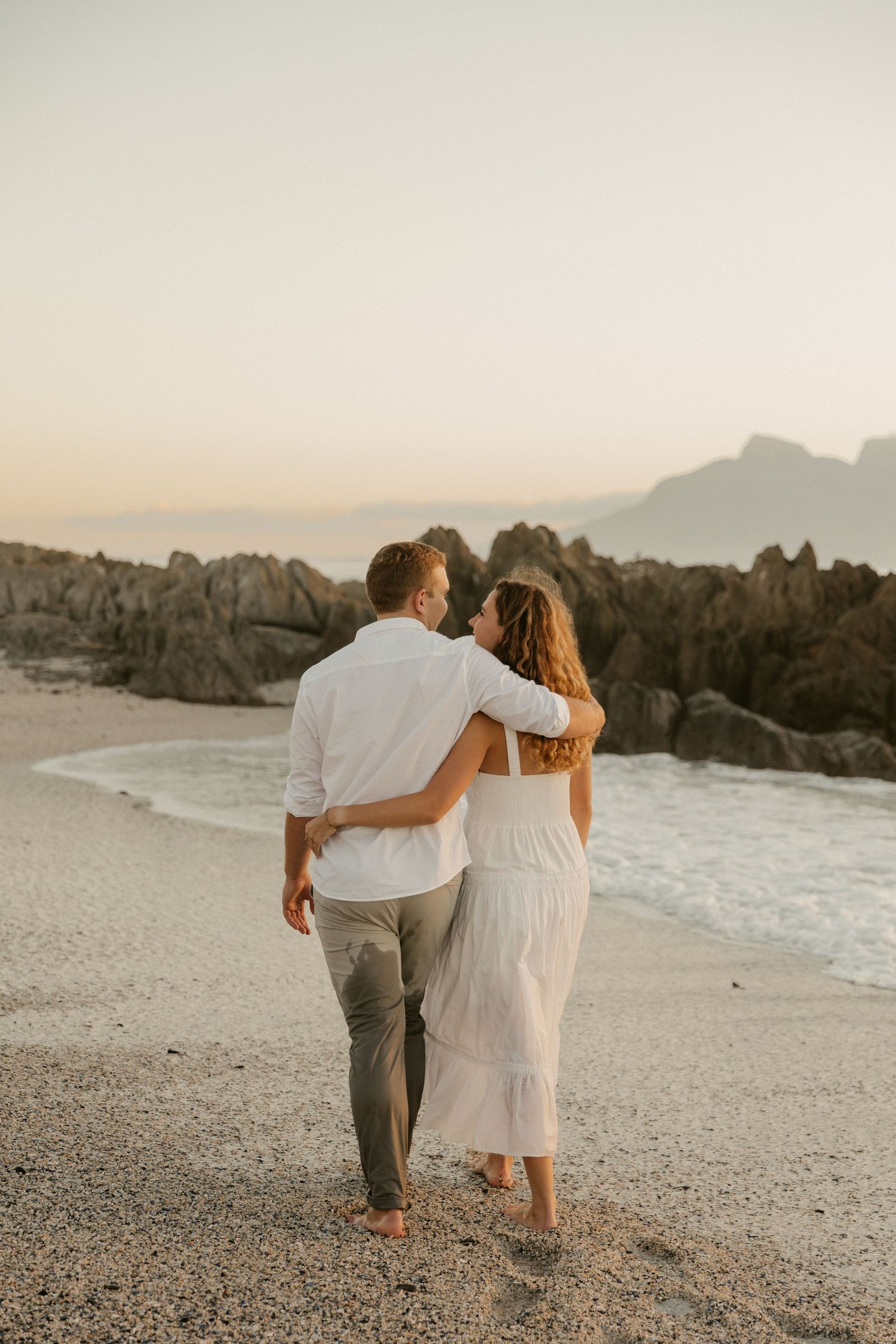 Couple walks arm in arm along a rocky beach during golden hour.