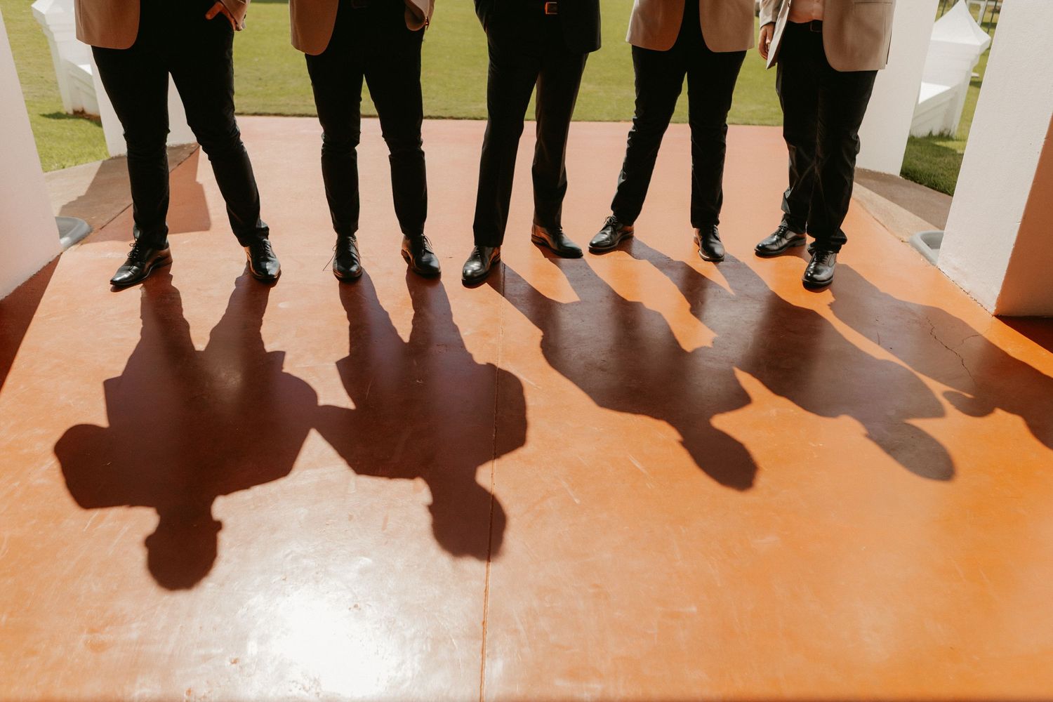 Groomsmen in black pants and dress shoes casting dramatic shadows on an orange floor at a wedding venue.