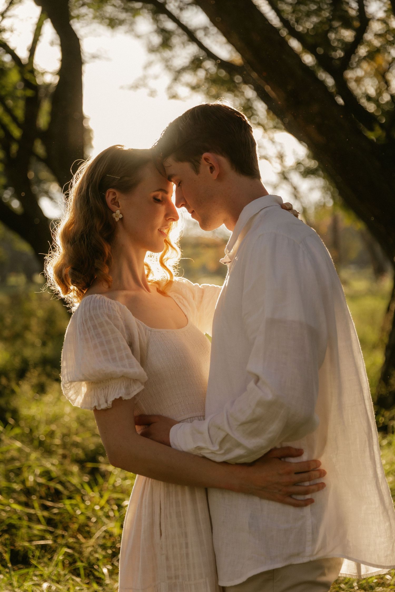 A romantic couple embraces in golden sunlight while standing in a lush outdoor natural setting during sunset.
