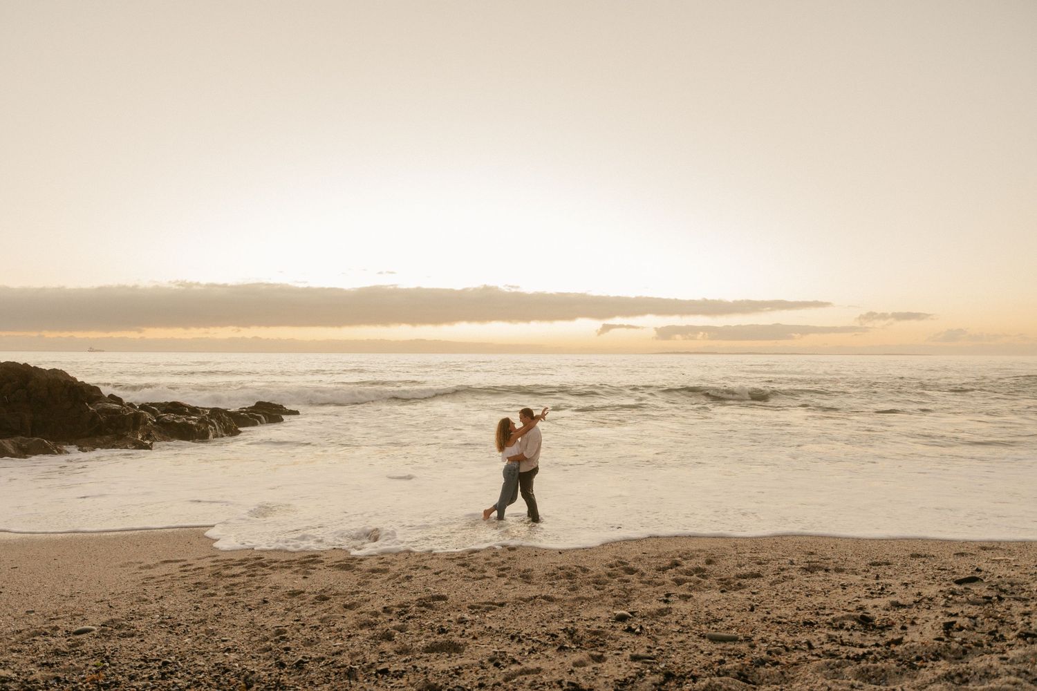 A romantic silhouette of a couple embracing on a beach at sunset with mountains in the distance.
