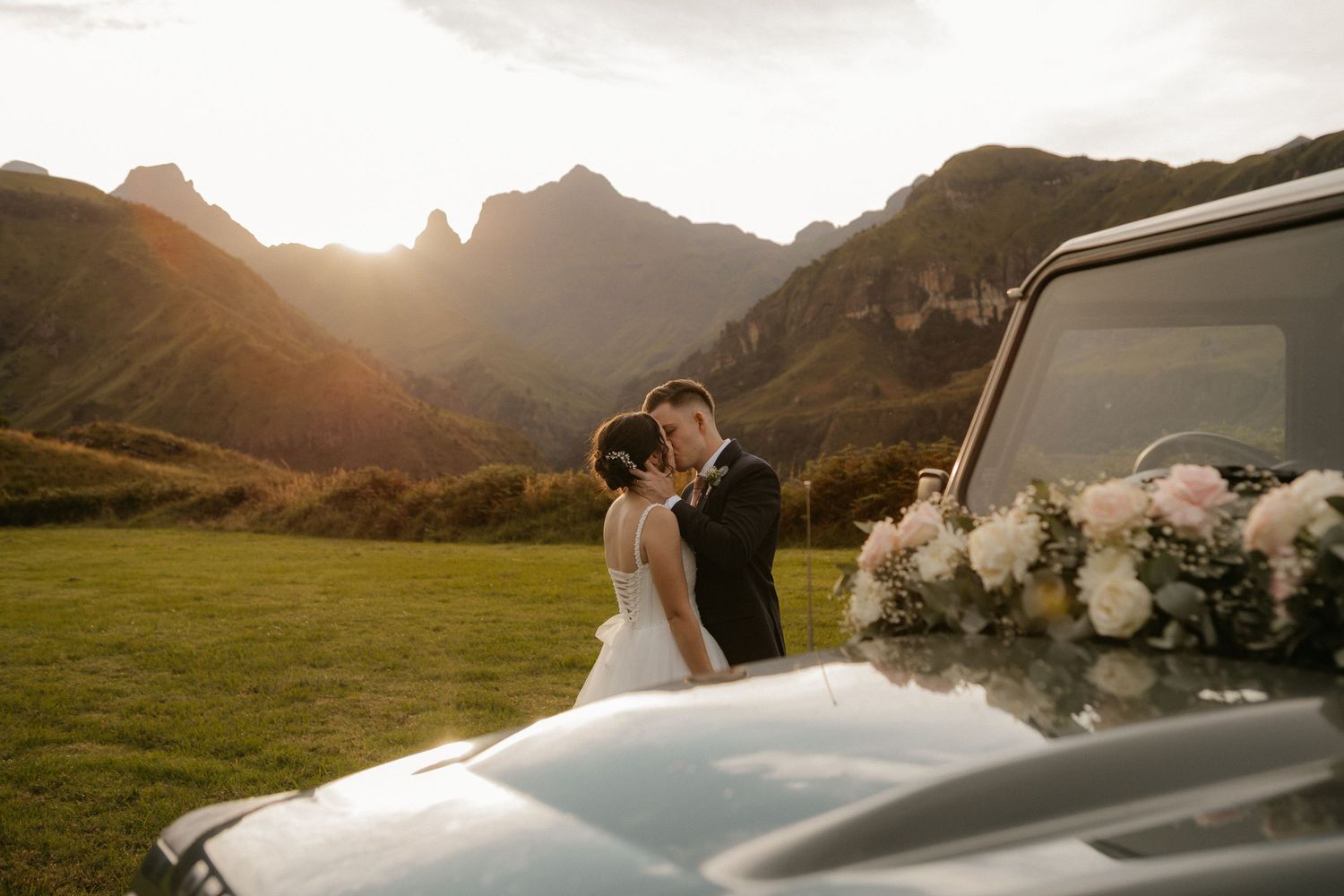 Bride and groom kissing at sunset beside a vintage car adorned with flowers, with dramatic mountain peaks in the background.