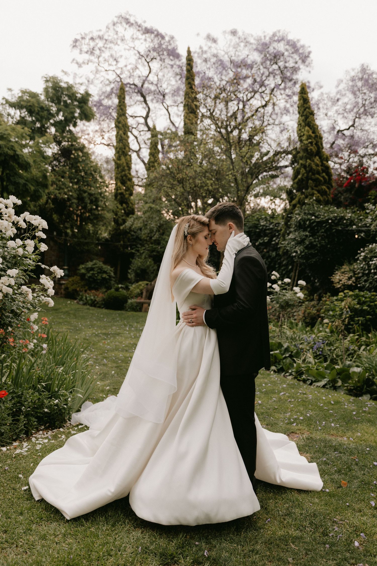 A romantic wedding couple embraces in a lush garden setting with tall cypress trees and blooming flowers.