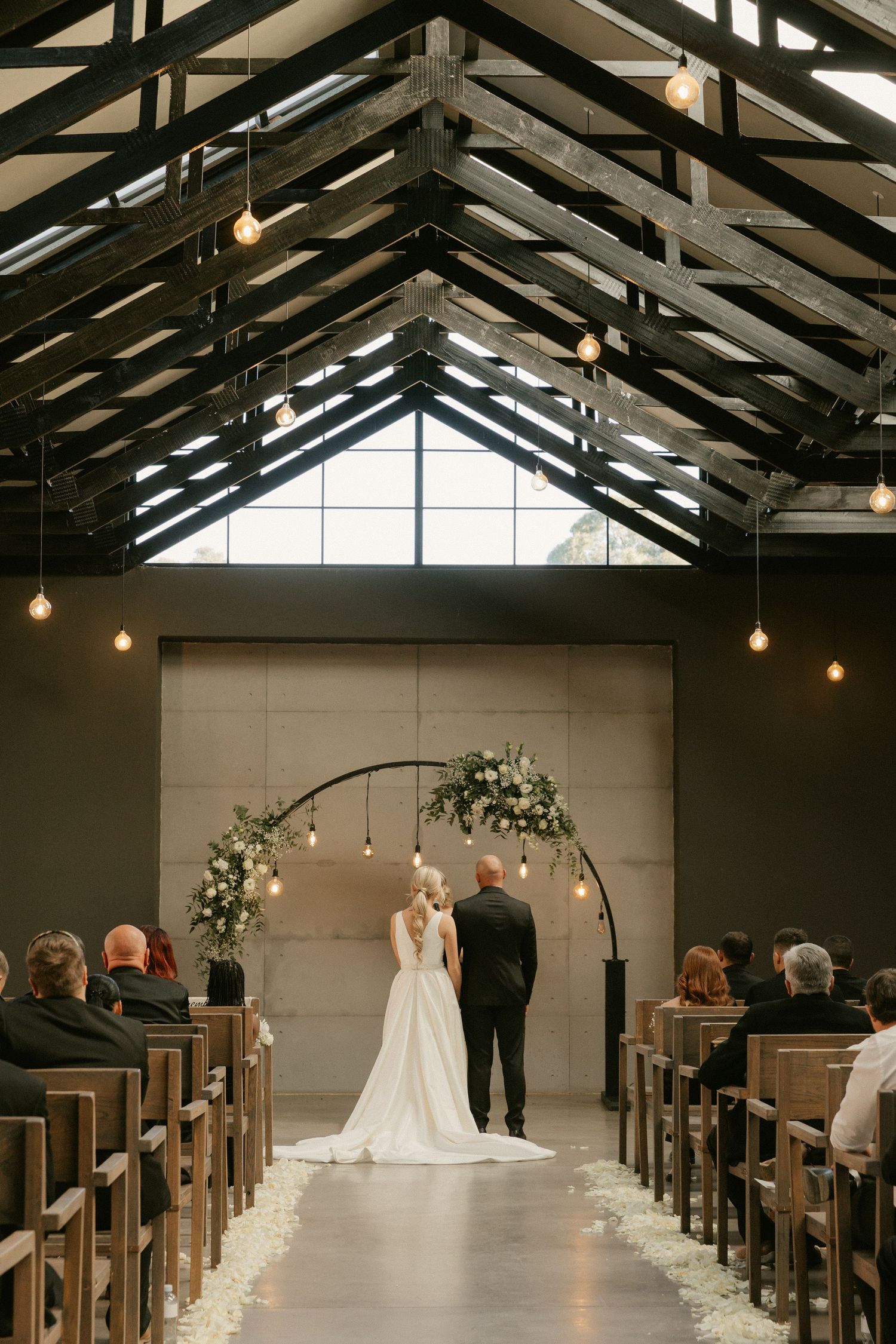 A church wedding ceremony in a modern chapel with exposed wooden beams, pendant lights, and wooden pews lining the aisle.