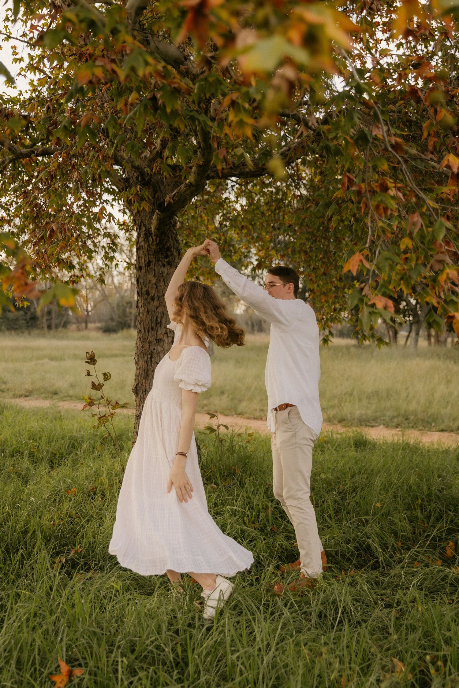 A couple dancing together under a tree with autumn leaves at golden hour in a rural field setting.