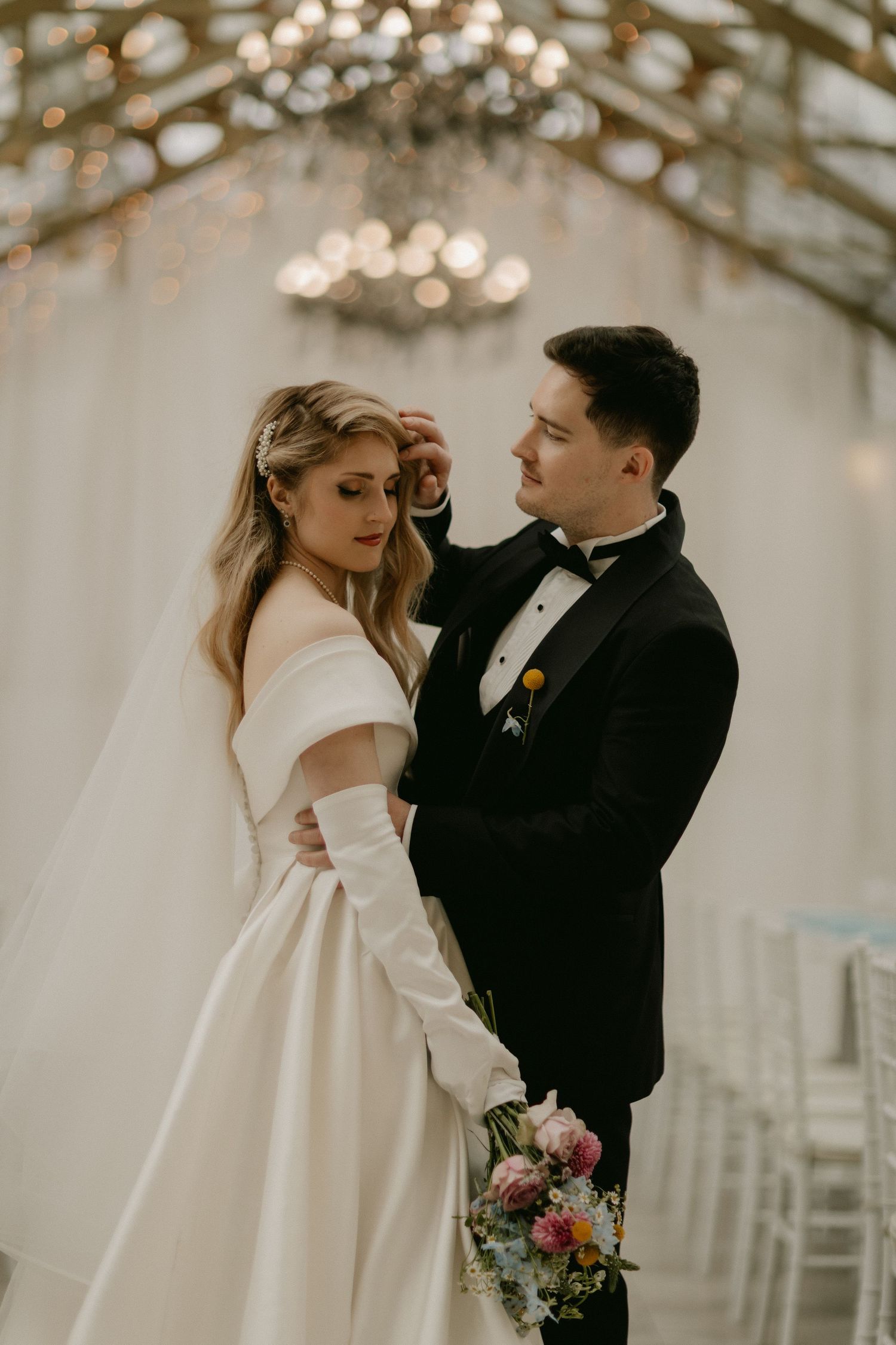 A romantic wedding couple embraces in an elegant indoor venue with twinkling lights and floral bouquet details.