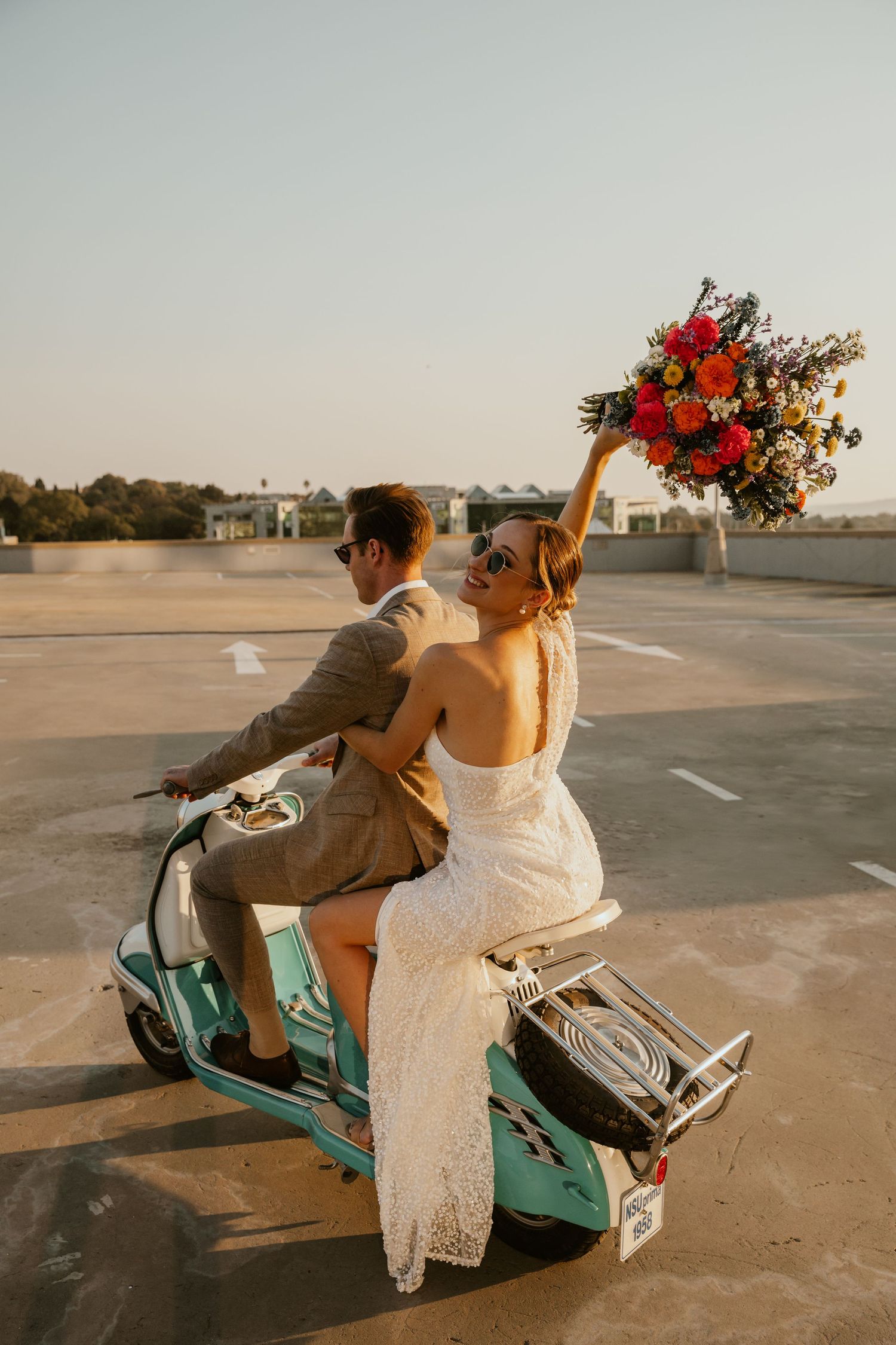 Newlyweds riding a mint green Vespa scooter at sunset.