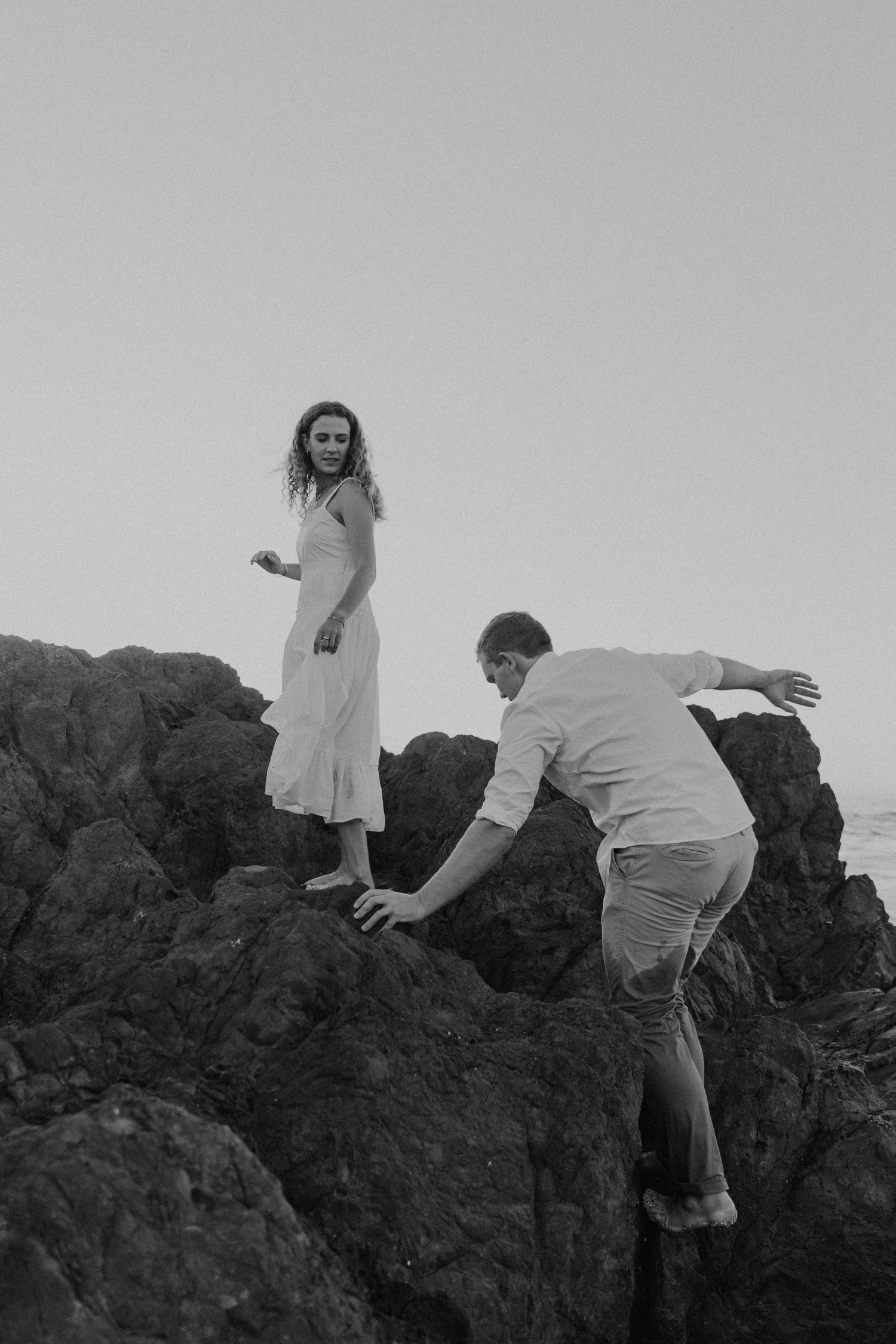Two people navigate rocky coastal terrain in black and white photography.