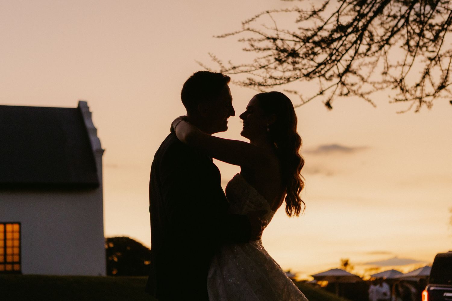 Silhouette of a couple embracing at sunset near a white chapel, with warm golden sky and bare tree branches.