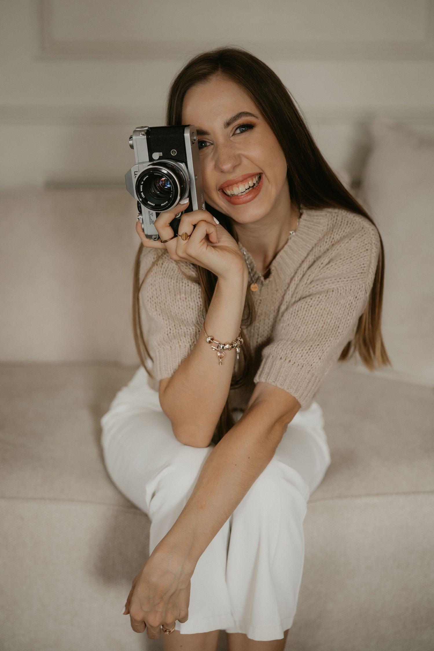 Smiling young woman holding a vintage camera while sitting on a couch in a beige knit top and white pants.