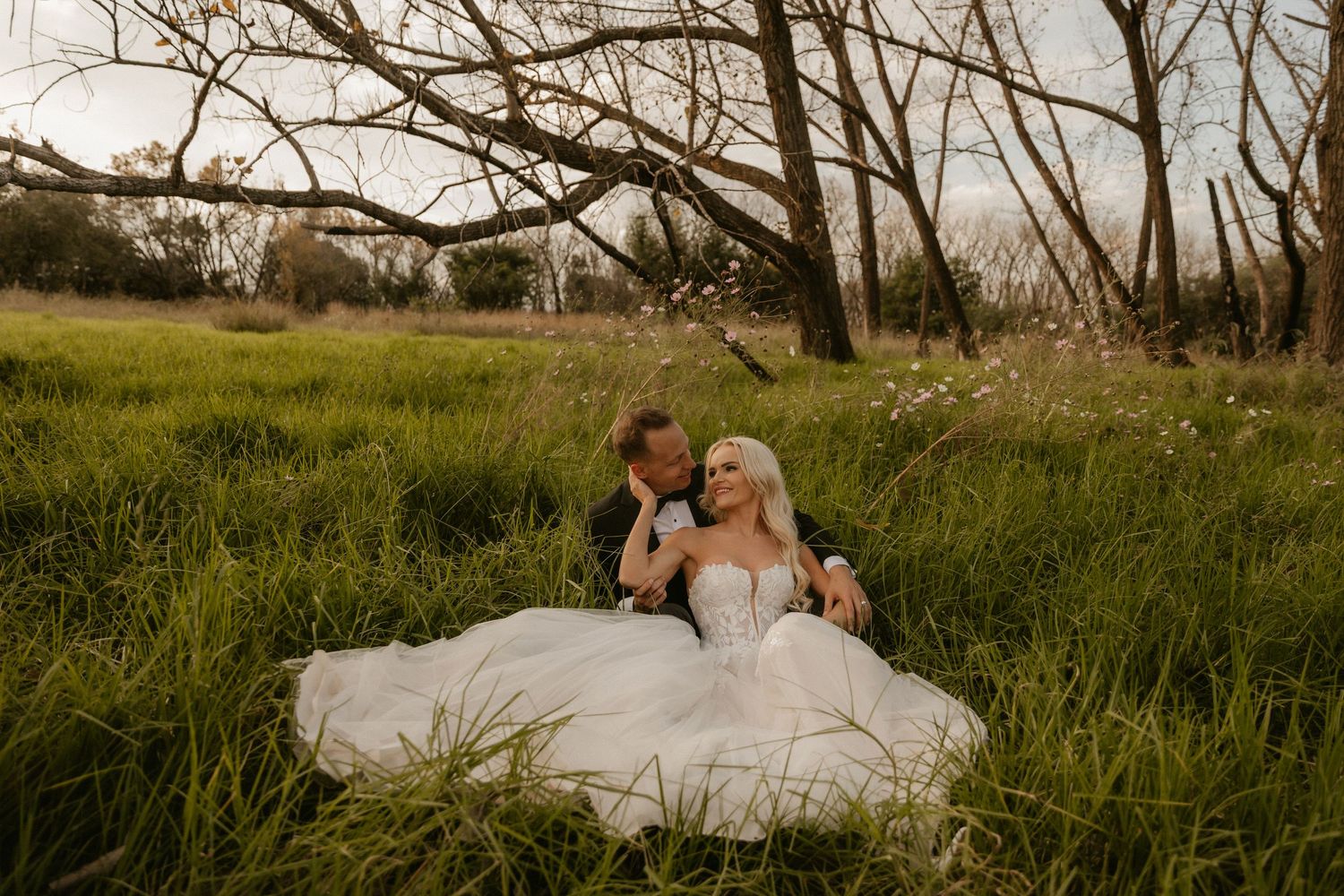Newlyweds relax together in tall grass during a sunset photoshoot in a rustic outdoor setting.