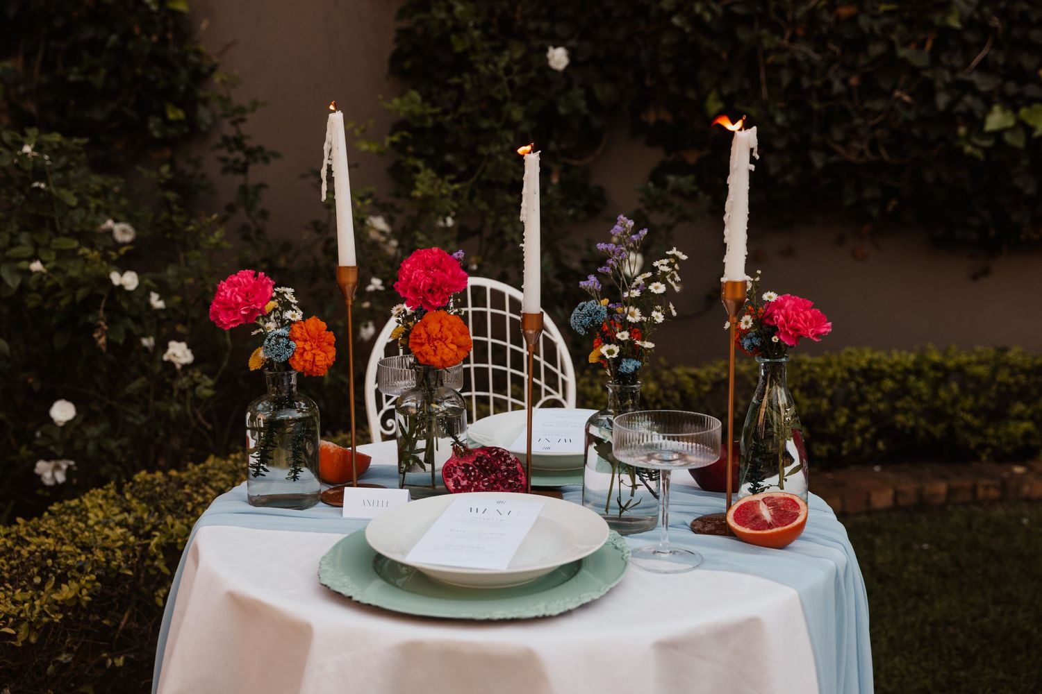 Intimate outdoor dining setup with white candles, flowers, and place settings.