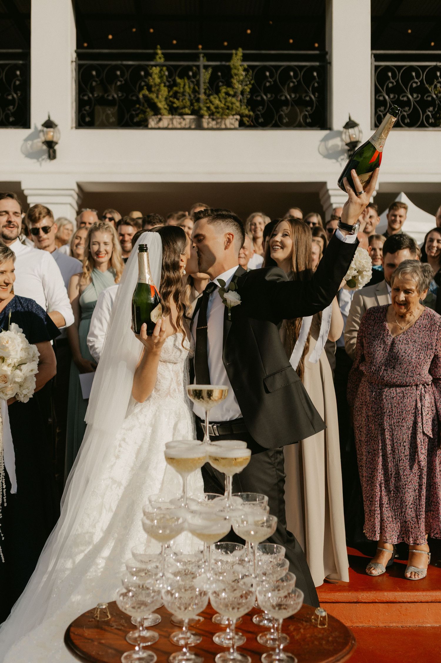Bride and groom celebrate with champagne tower surrounded by cheering wedding guests.