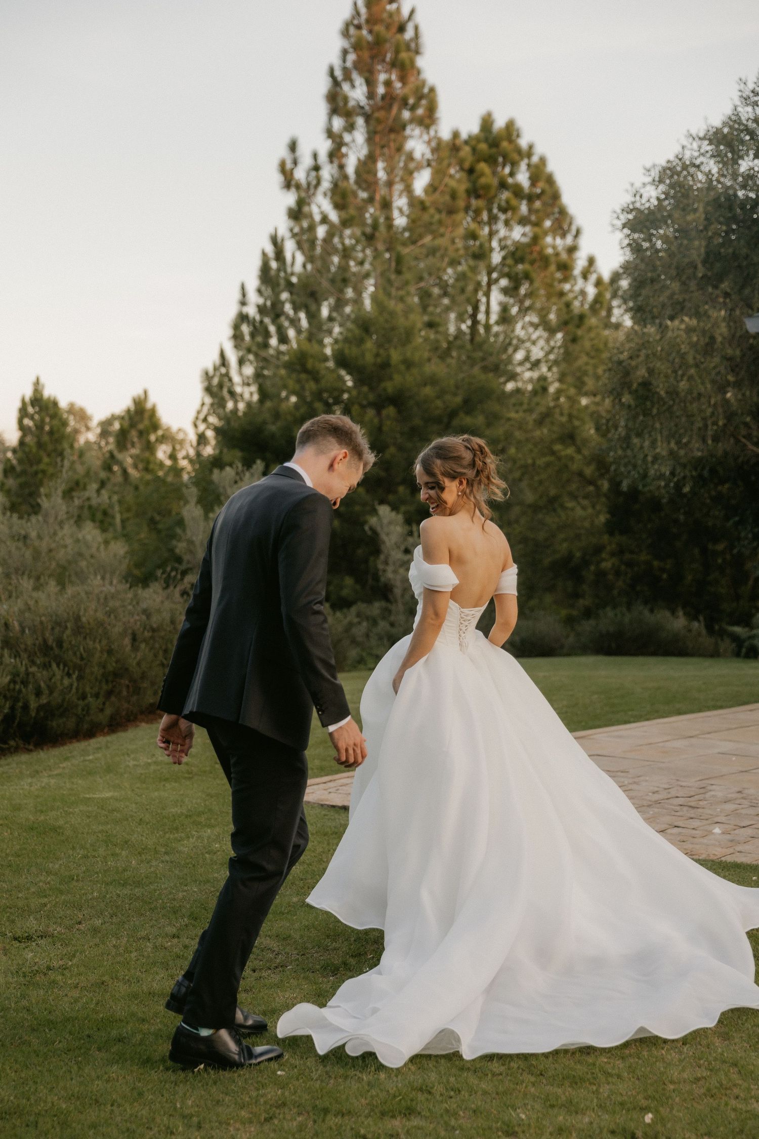 A couple in wedding attire walks together on a path surrounded by green trees and grass during golden hour.