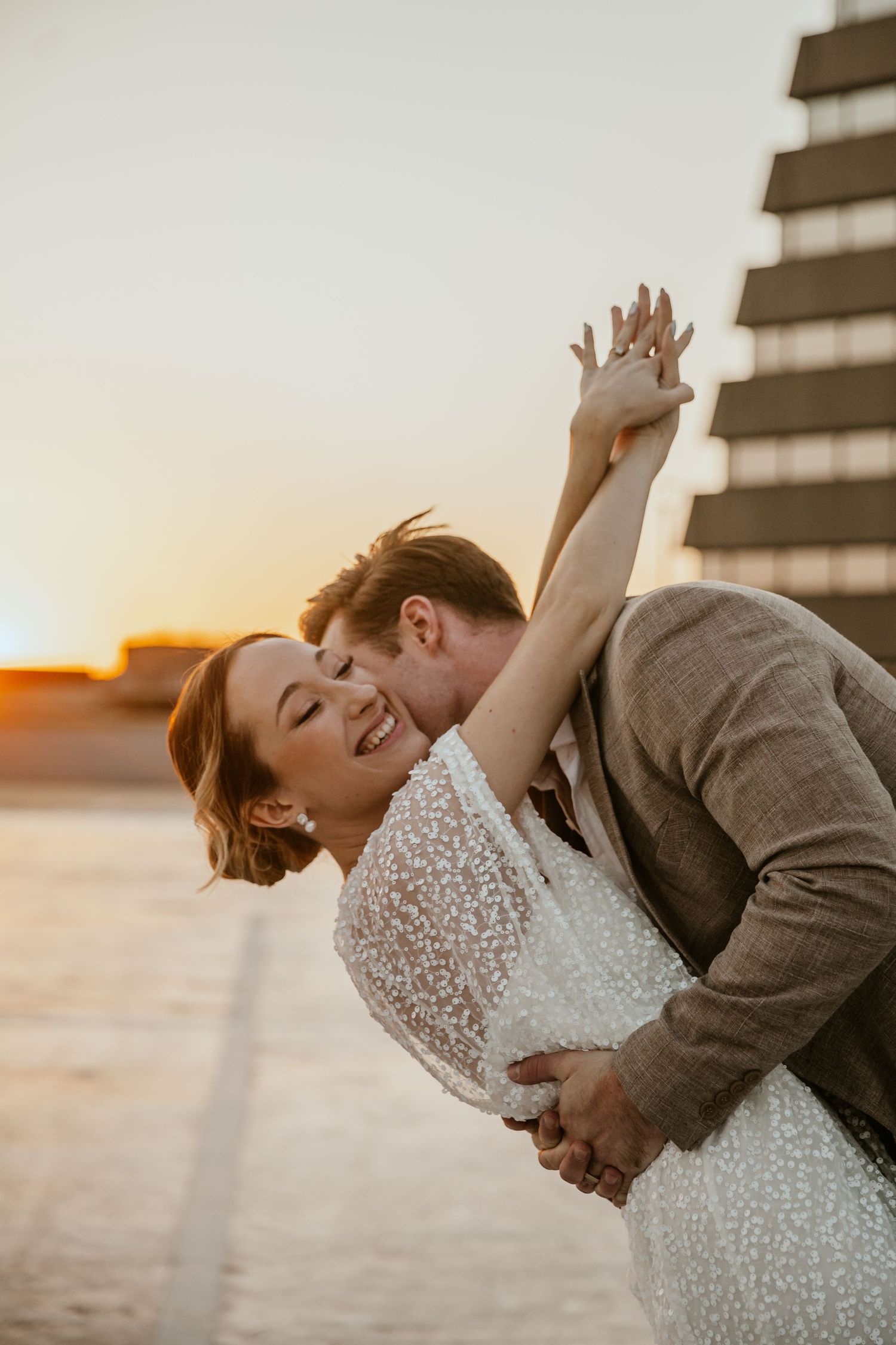 Romantic couple embracing during golden hour sunset next to modern building.