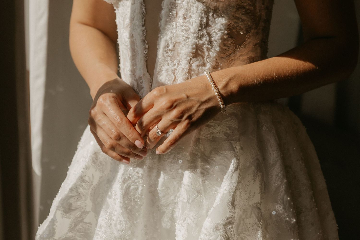 Close-up of bride in lace wedding gown with pearl bracelet and ring, hands clasped at waist.