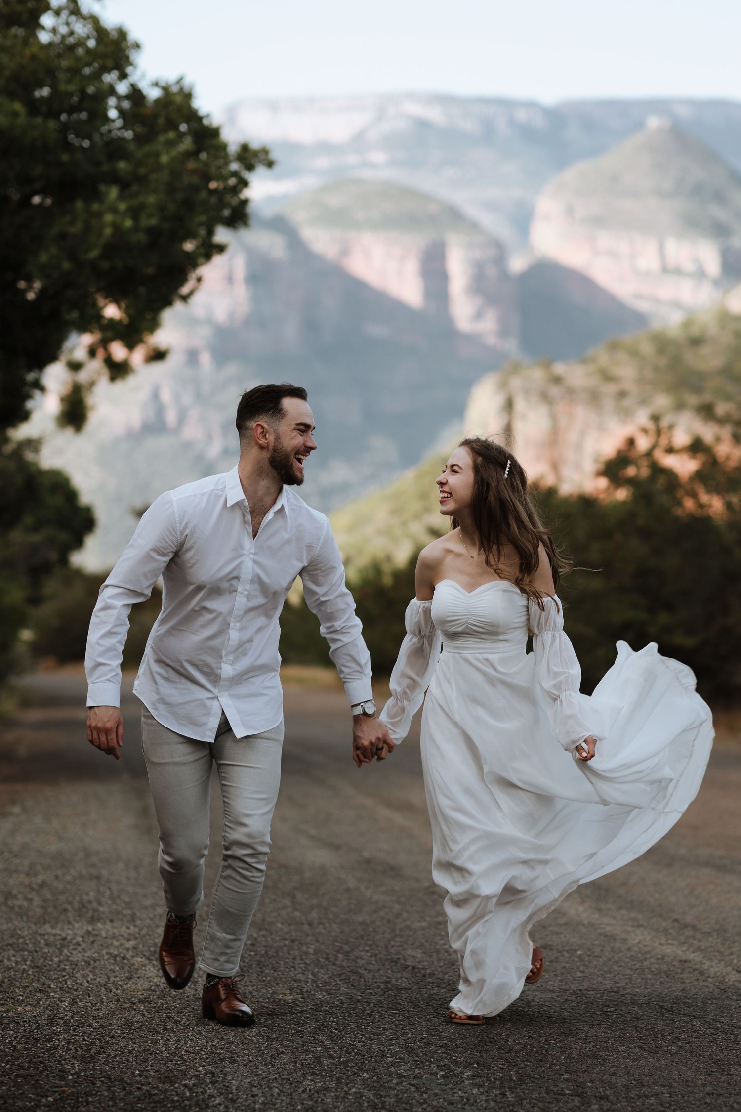 Couple walking hand in hand on mountain path with flowing white dress and dramatic cliff backdrop.