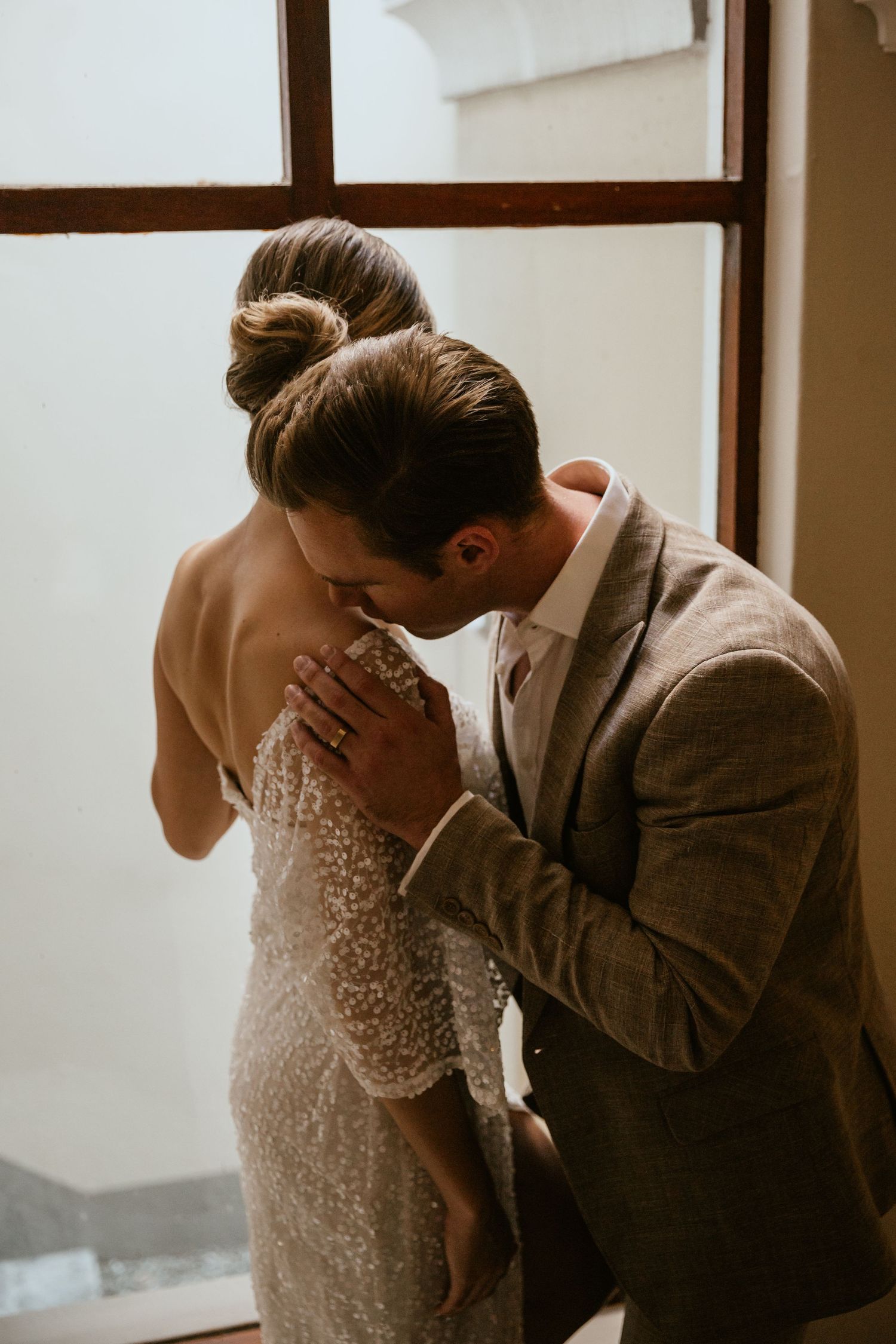 A tender moment between newlyweds sharing an intimate embrace by a window in soft lighting.