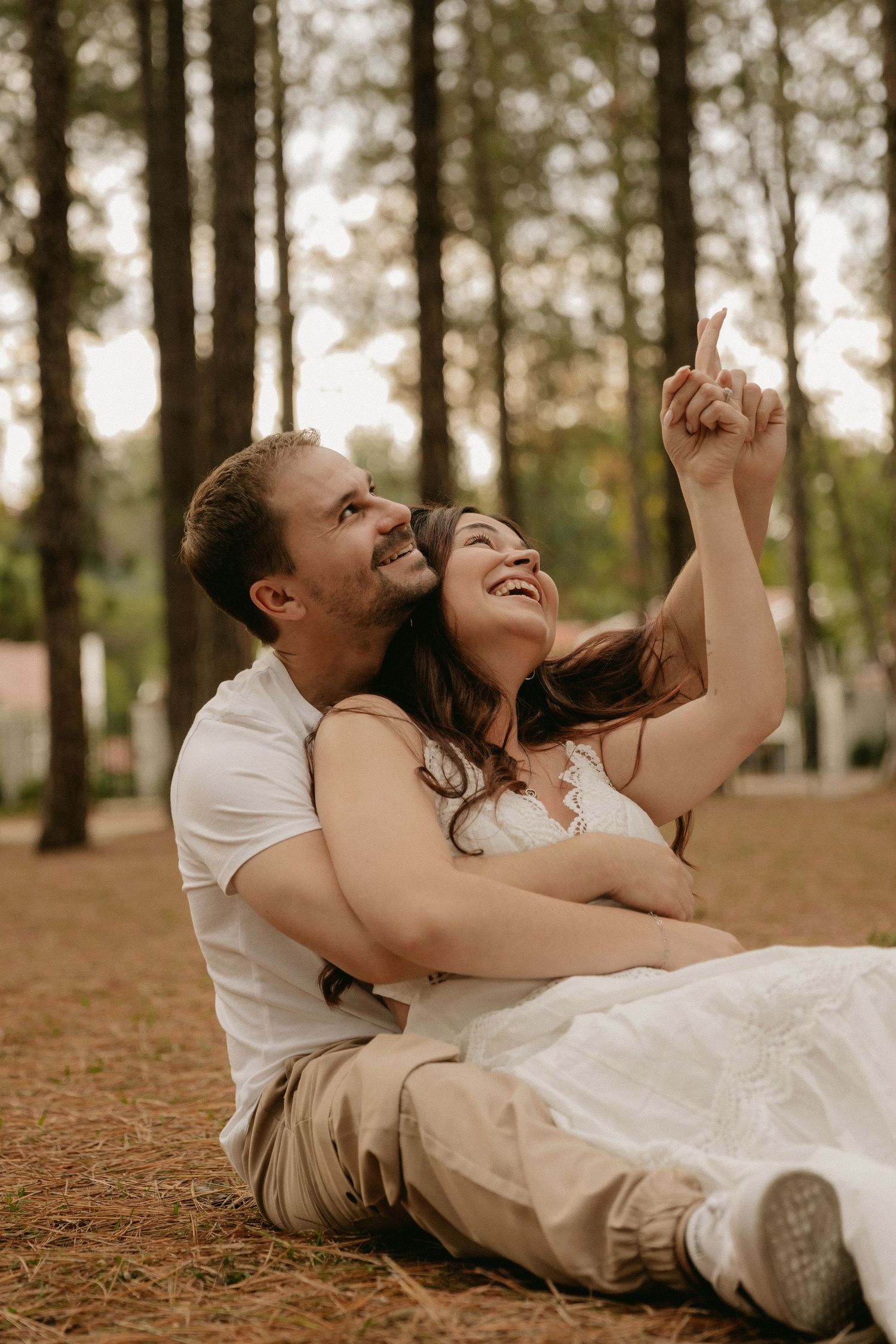 A couple in white clothing lies on autumn ground in a pine forest, laughing joyfully together in a candid moment.