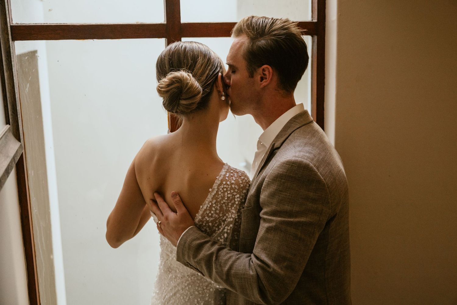 Romantic wedding moment captured by window light with couple in intimate embrace.