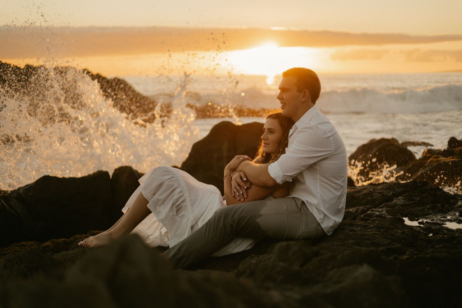 Two figures embrace on rocky coastal cliffs at sunset while ocean waves crash in the background.