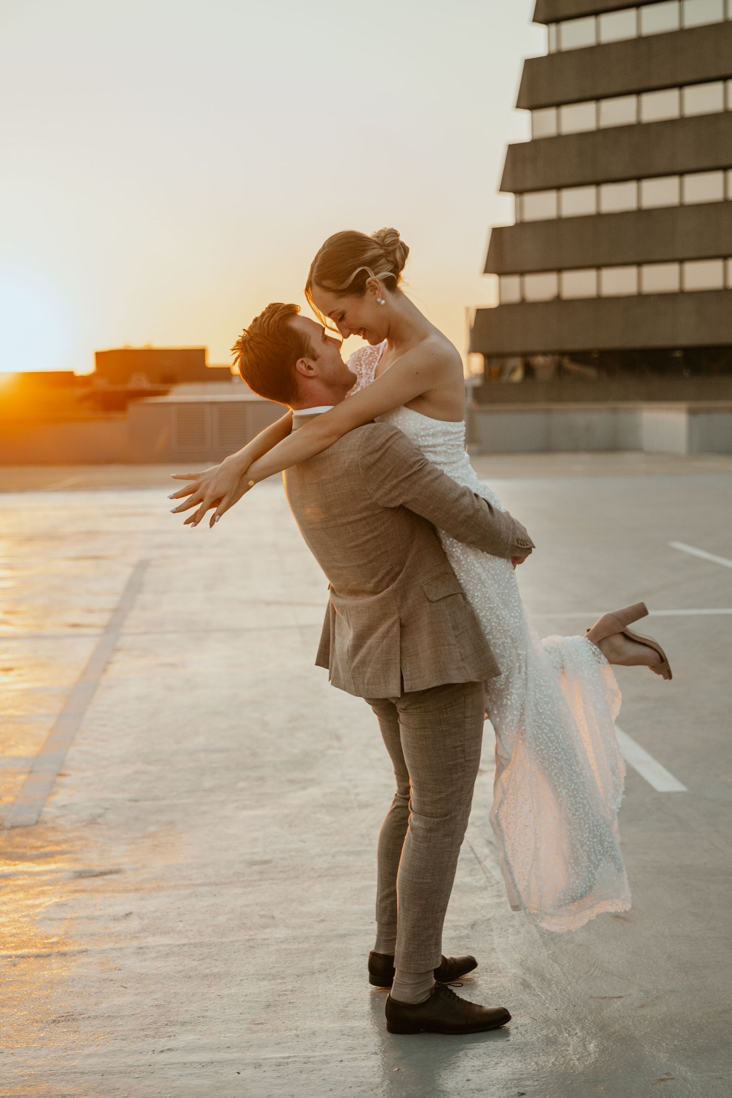A romantic sunset rooftop moment as a couple embraces in formal wedding attire.