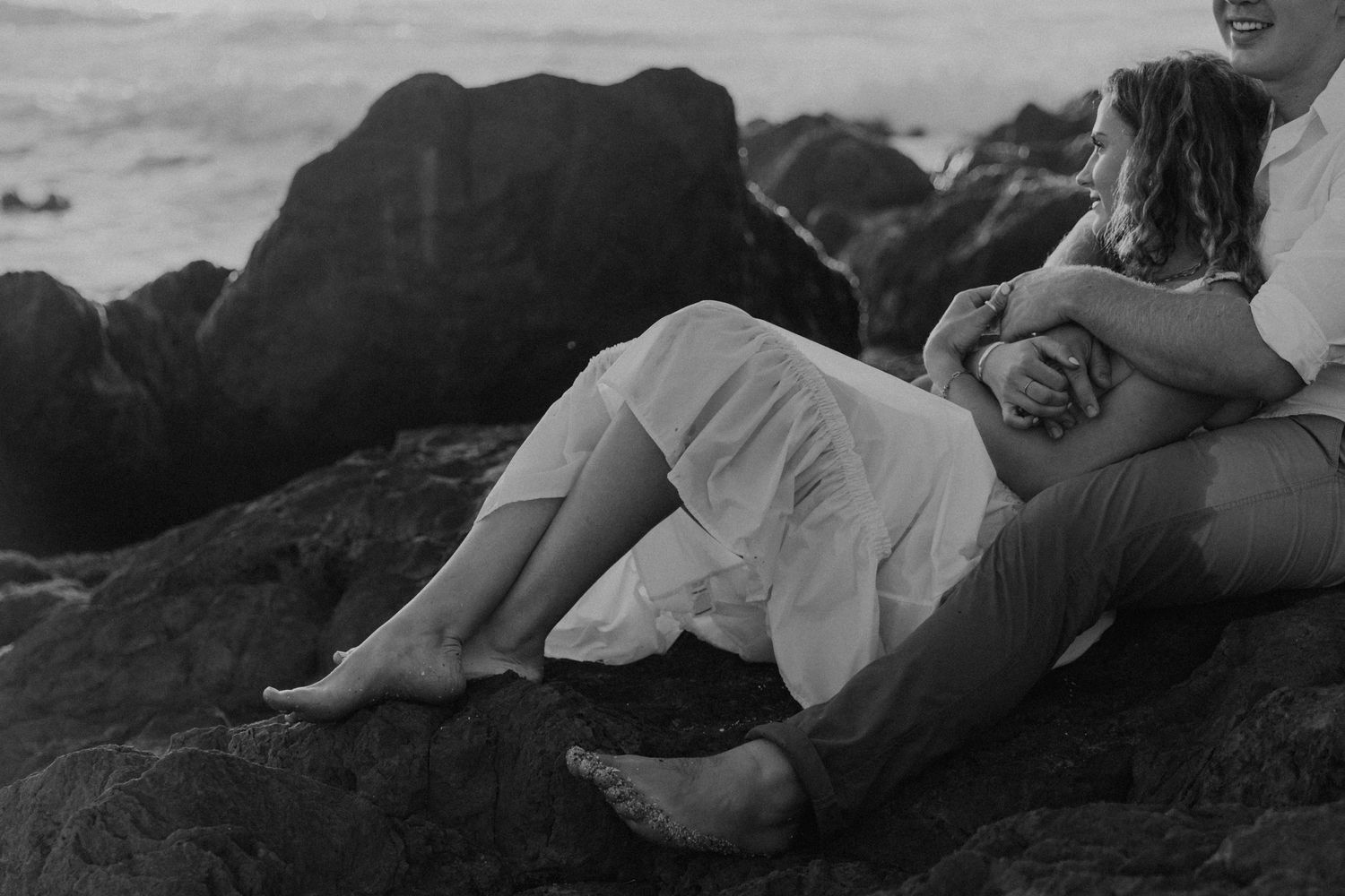 A black and white photo captures an intimate moment on coastal rocks at sunset.