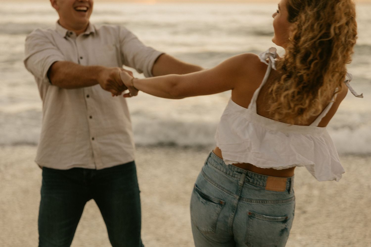 Couple dances playfully on the beach at golden hour with the ocean behind them.