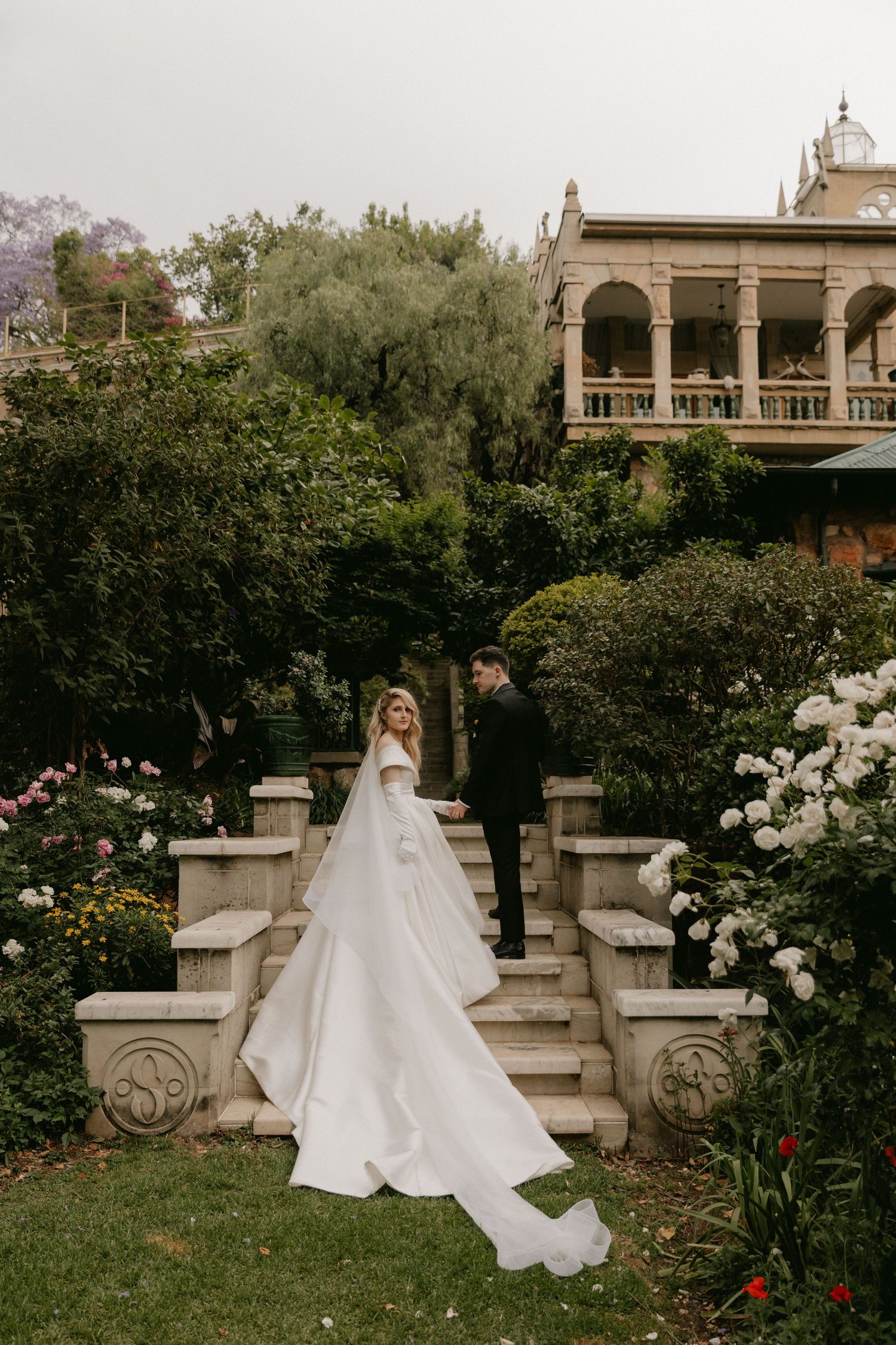 A bride in a flowing white ballgown poses elegantly in a lush garden setting with classical architecture.