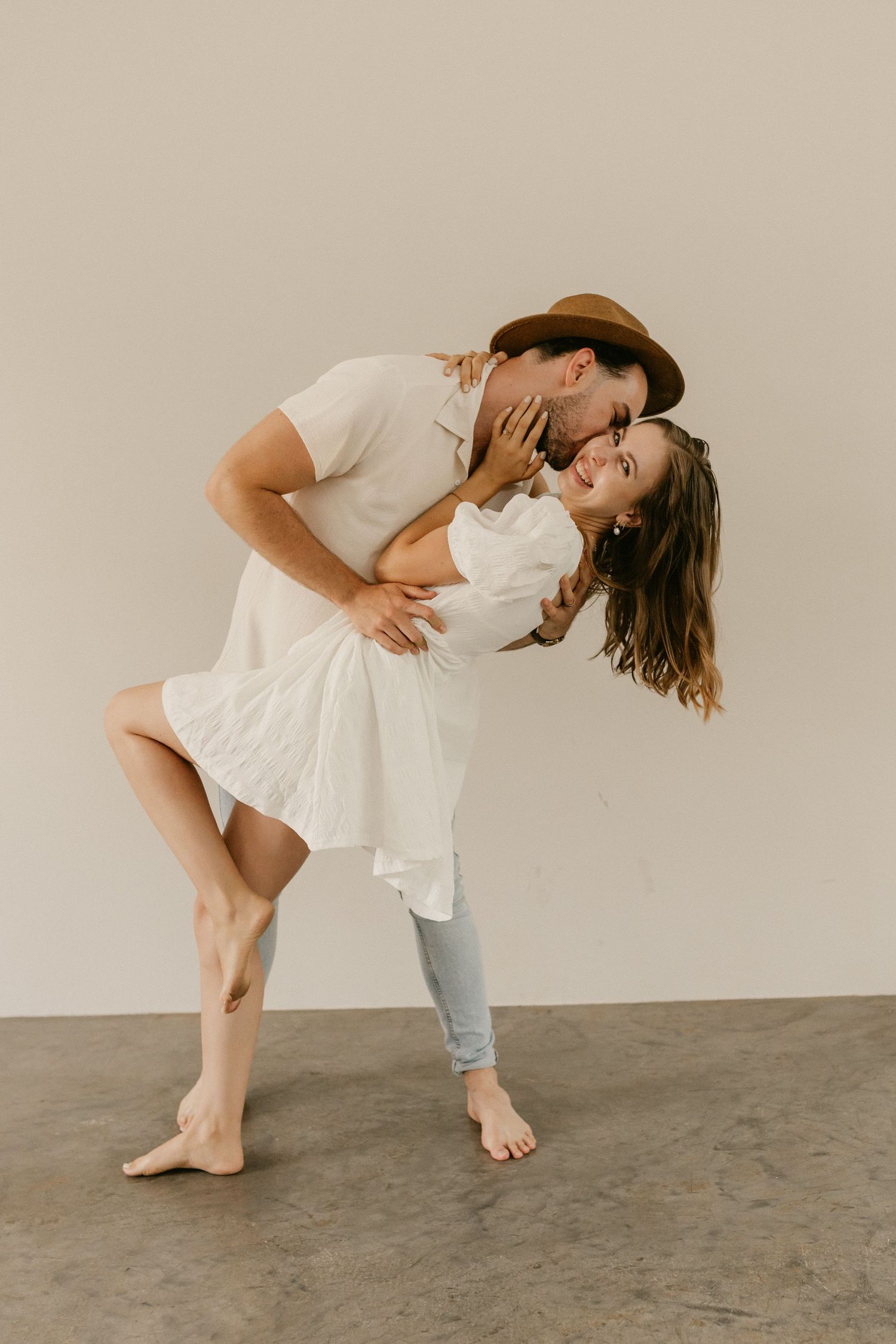 A romantic dance pose with a barefoot couple, one in a white dress and one in jeans and a hat, on a gray floor.