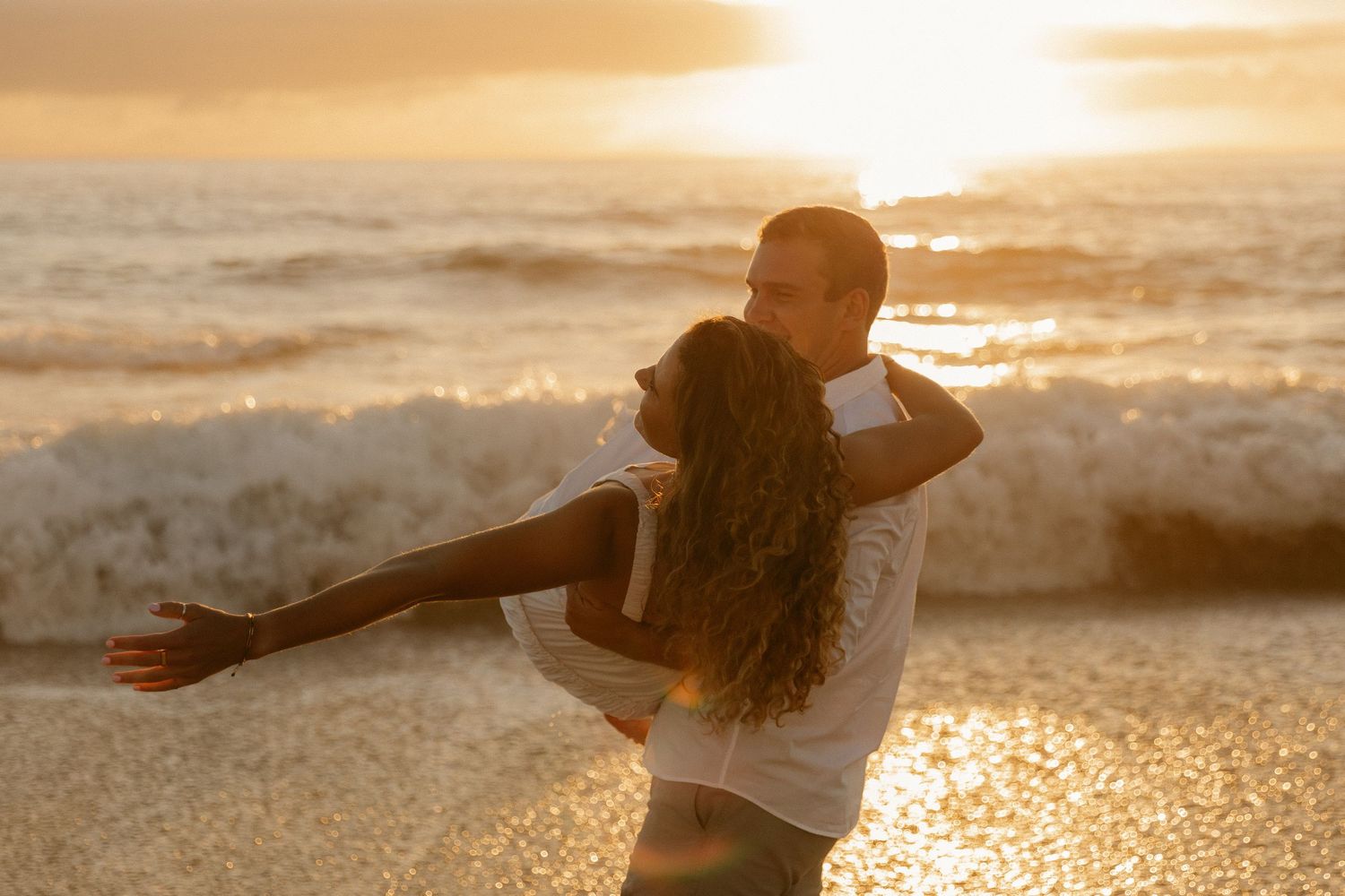 A couple shares an intimate moment on a beach as golden sunlight illuminates crashing waves.