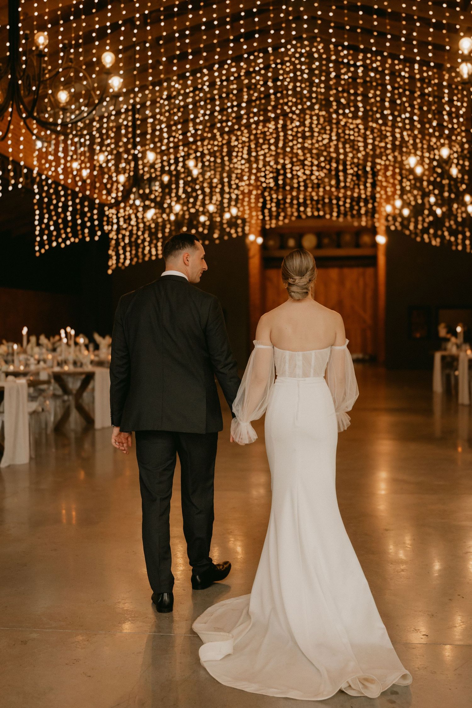 A couple walks under a stunning ceiling of warm glowing string lights at an elegant wedding reception venue.