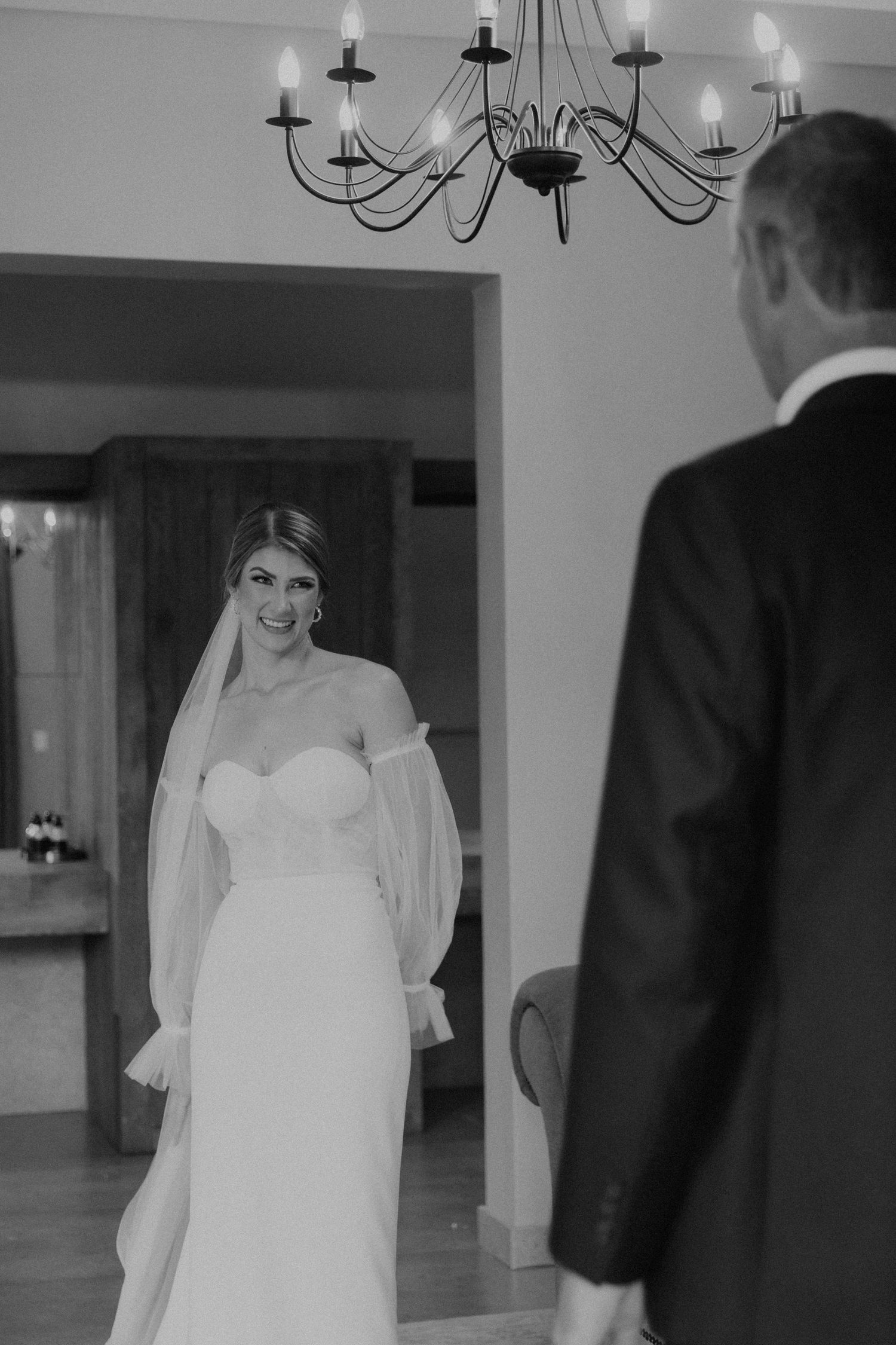 Black and white wedding photo showing bride in white strapless gown and veil with chandelier overhead.