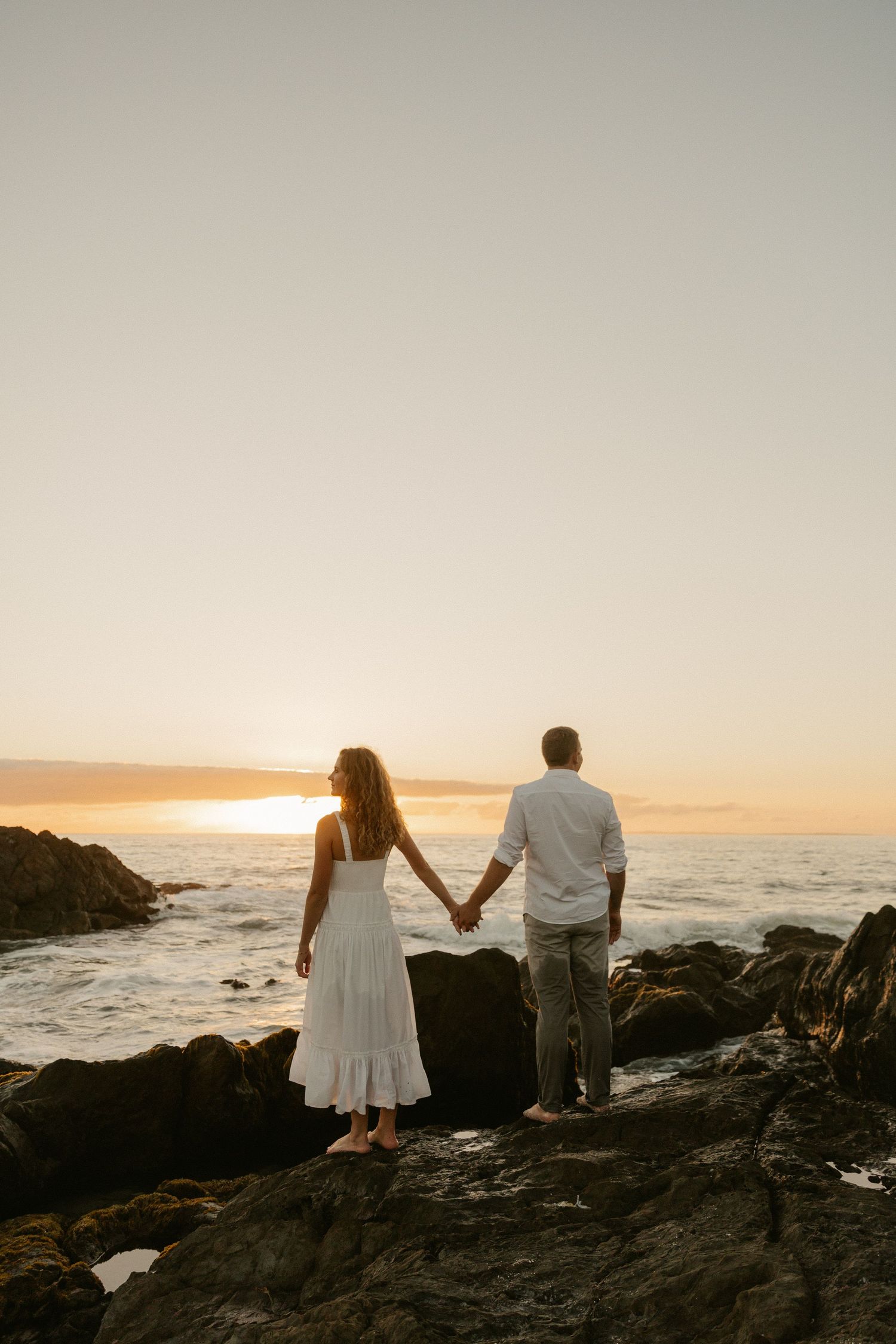 A couple stands hand in hand on rocky coastline overlooking ocean at sunset.