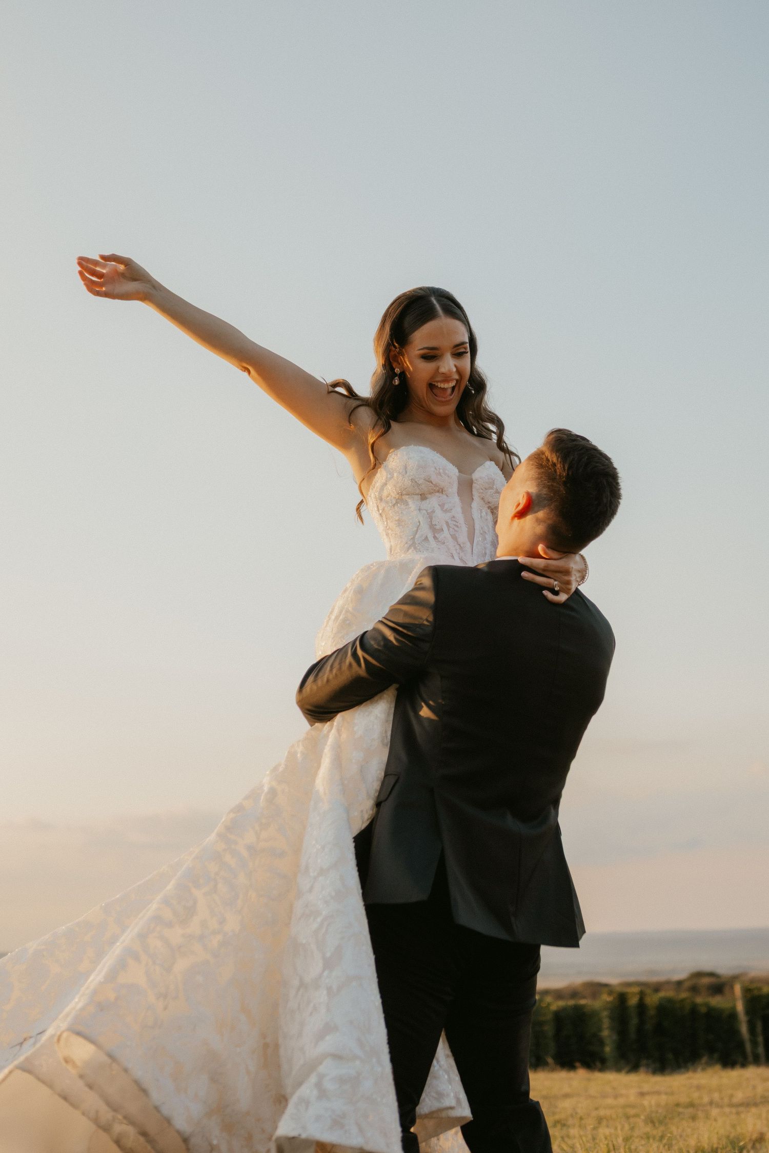 Groom lifts smiling bride in white lace gown at golden hour, her arm outstretched joyfully against a clear sky.